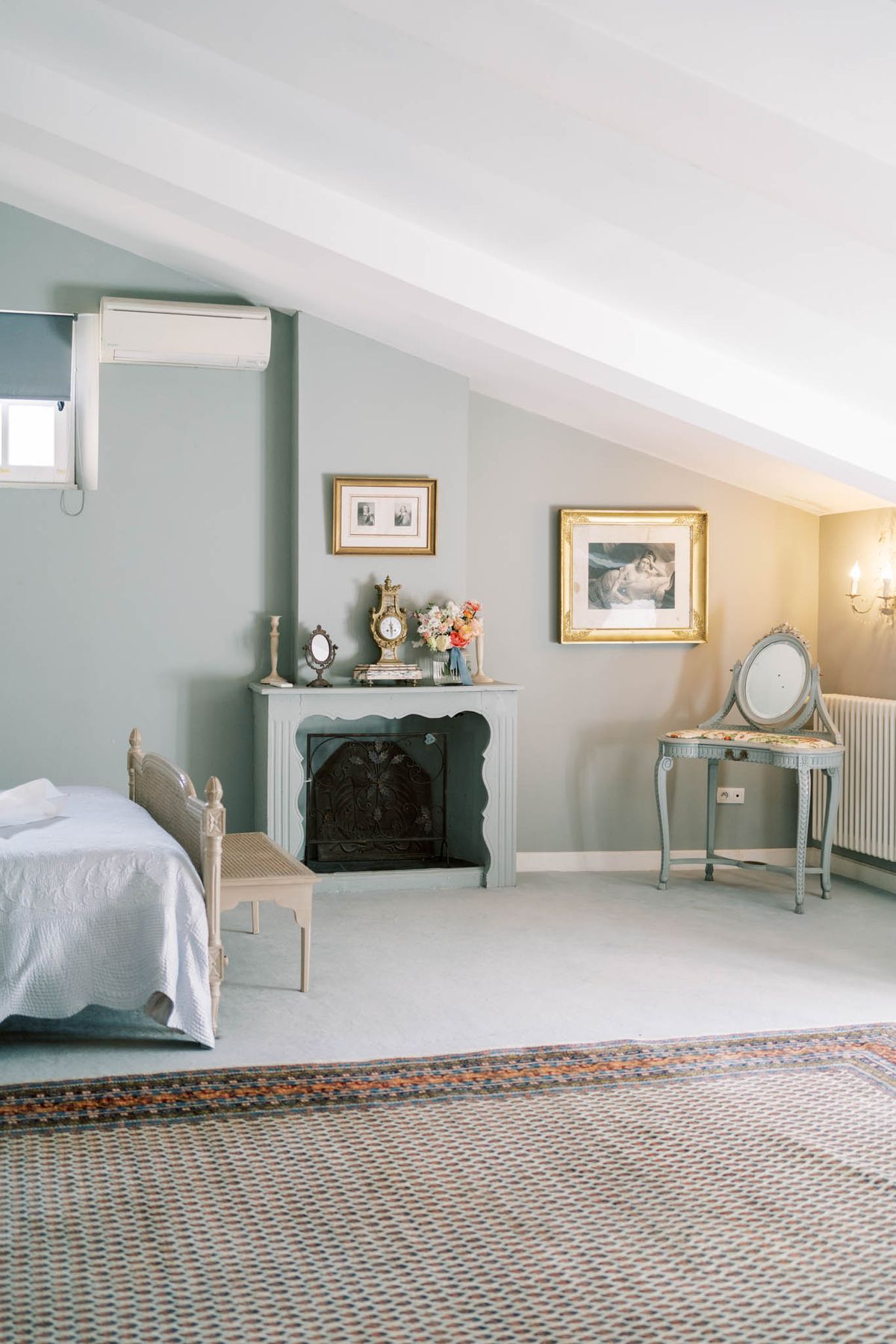 An interior room shot of a getting-ready suite, likely within a château or manor house venue. The room features sloped attic-style ceilings, sage-green painted walls, and a decorative white fireplace mantel dressed with a small bouquet of coral and peach flowers, a gilt clock, a candlestick, and a vanity mirror. Furnishings include a single bed with a white quilted cover and a painted wood cane bench at its foot, a pale blue Louis XVI-style vanity chair with a circular mirror, and a patterned area rug in terracotta and blue tones. Two gold-framed artworks hang on the walls, and a wall sconce provides warm lighting on the right side. The composition is a wide interior shot with no people present. Potential venue feature image.