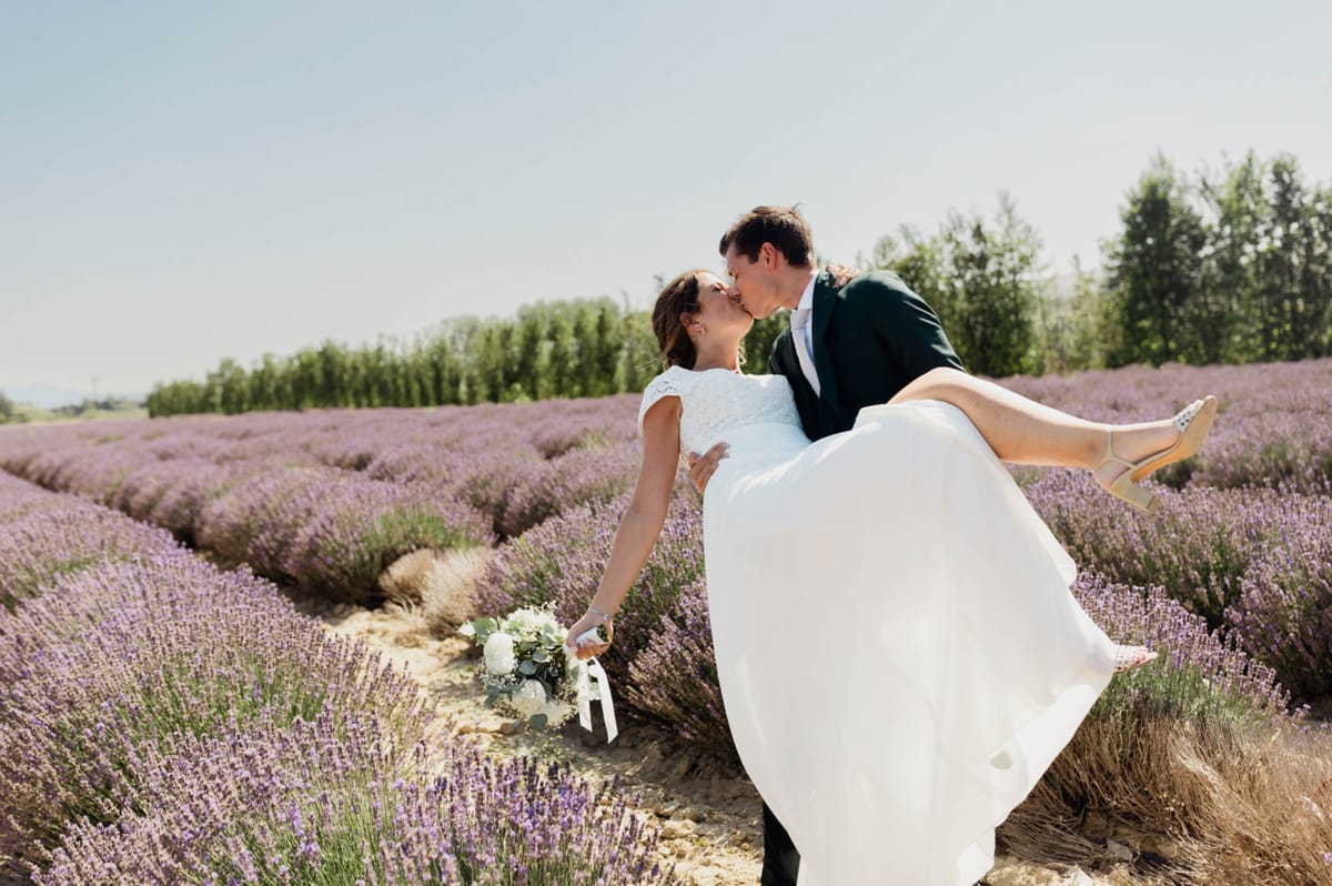 A couple portrait shot outdoors in a lavender field in full bloom, with rows of purple lavender extending into the background. The groom, wearing a dark forest green suit, is lifting and kissing the bride, who wears a white cap-sleeve lace-bodice gown with a flowing skirt and gold strappy heels. The bride holds a bouquet of white peonies or ranunculus with eucalyptus greenery and white ribbon trails. The composition is a mid-range portrait taken at ground level, with the lavender rows creating natural leading lines behind the couple.