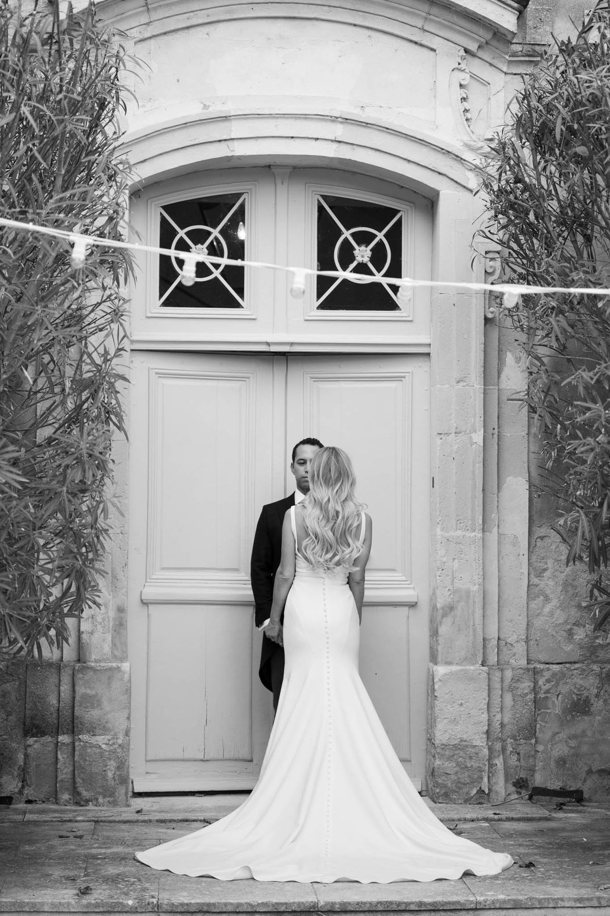 A black-and-white couple portrait taken outdoors in front of a large arched double door set into a stone-cut facade, consistent with a French château or manor house. The bride and groom stand facing each other and holding hands, photographed from behind; the bride wears a fitted mermaid-style gown with a low open back, button detail running down the spine, thin straps, and a floor-length train spreading across the stone paving. Her long, wavy blonde hair falls down her back. The groom is dressed in a dark suit. A string of festoon lights is visible crossing the frame horizontally in the upper portion of the image. The shot is a full-length portrait with moderate contrast, capturing the architectural detail of the ornate door surround. Potential venue feature image.