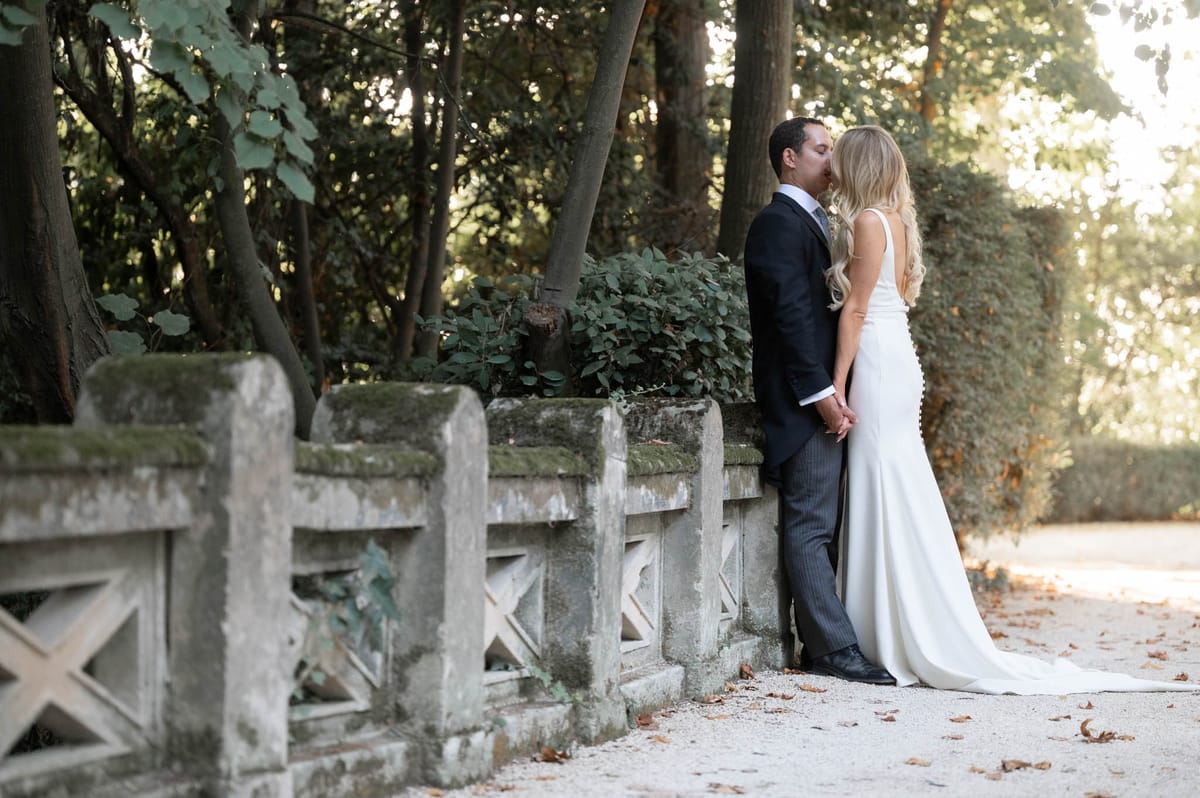 Couple kisses beside ornamental stone balustrade on gravel path with trimmed hedgerows and tall trees