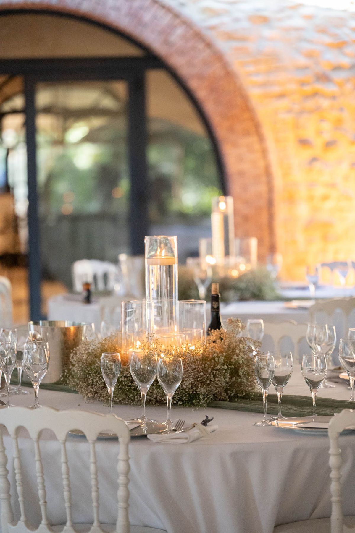 A close-up detail shot of a wedding reception table setting inside a venue with exposed brick arched walls and large arched glass doors. The round table is dressed in a white linen tablecloth and set with white Chiavari chairs, clear wine glasses, and silver cutlery on white napkins. The centerpiece features graduated clear cylinder vases with floating candles surrounded by a bed of dried gypsophila (baby's breath) and a dark green leaf runner, creating a warm candlelit glow. Additional tables with candelabras and similar floral arrangements are visible in the soft background, suggesting a classic, candle-heavy decor palette in white, gold, and green.