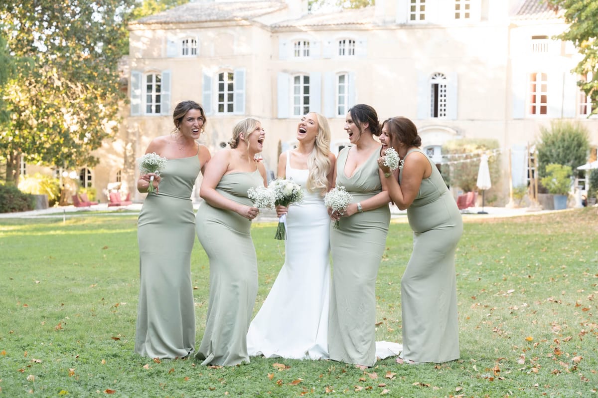 Bride and four bridesmaids in sage green slip dresses holding baby's breath bouquets laughing before chateau