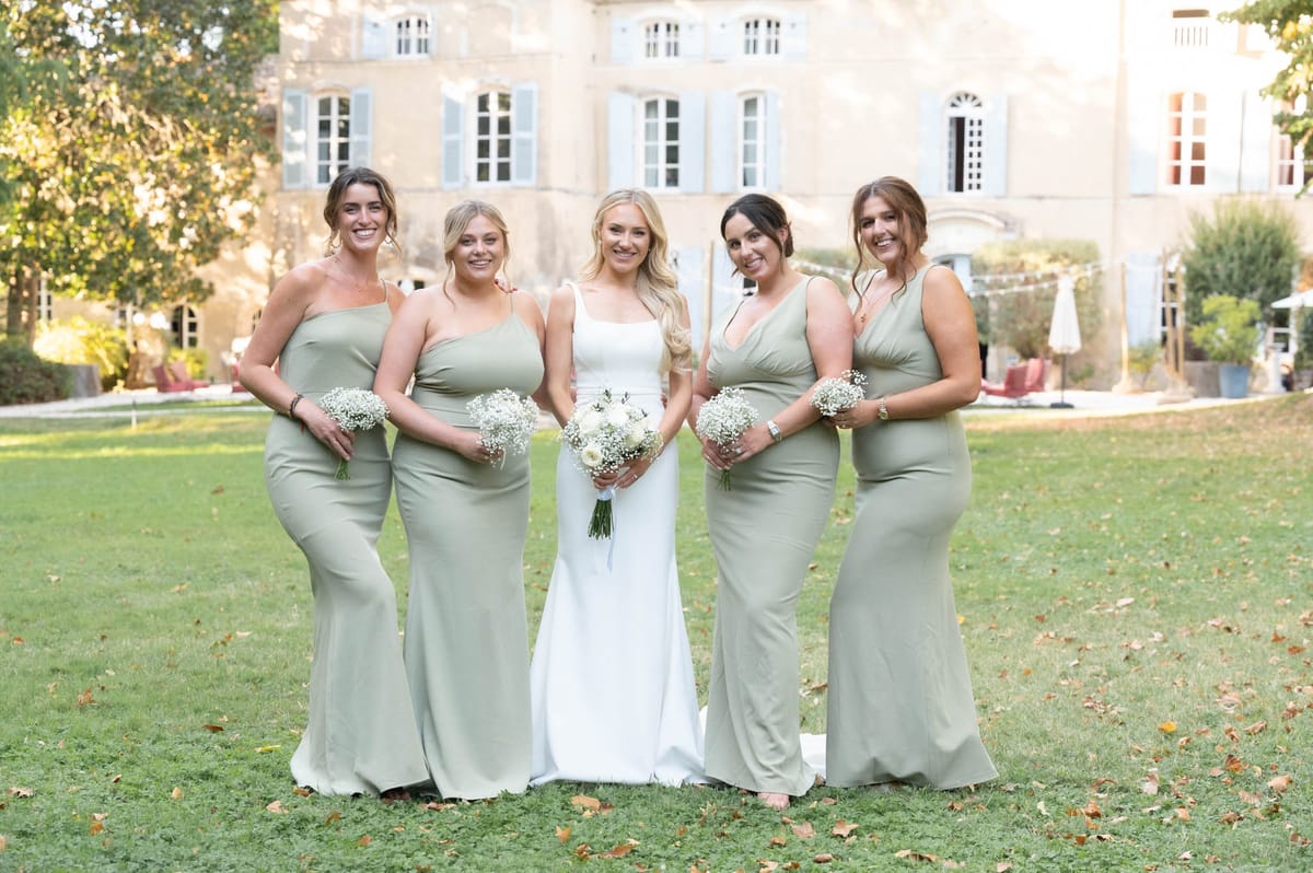 A bridal party portrait taken outdoors on the grounds of a French château, with the pale stone façade and pale blue shutters visible in the background. The bride stands in the center wearing a sleek, square-neckline white gown with thin straps, holding a bouquet of white ranunculus and greenery. She is flanked by four bridesmaids in floor-length sage green fitted dresses in varying neckline styles — one-shoulder, V-neck, and sleeveless — each holding a small bunch of white gypsophila. The overall color palette is white and sage green, giving a clean, understated aesthetic. This is a medium wide-shot group portrait with all five women facing the camera and smiling.