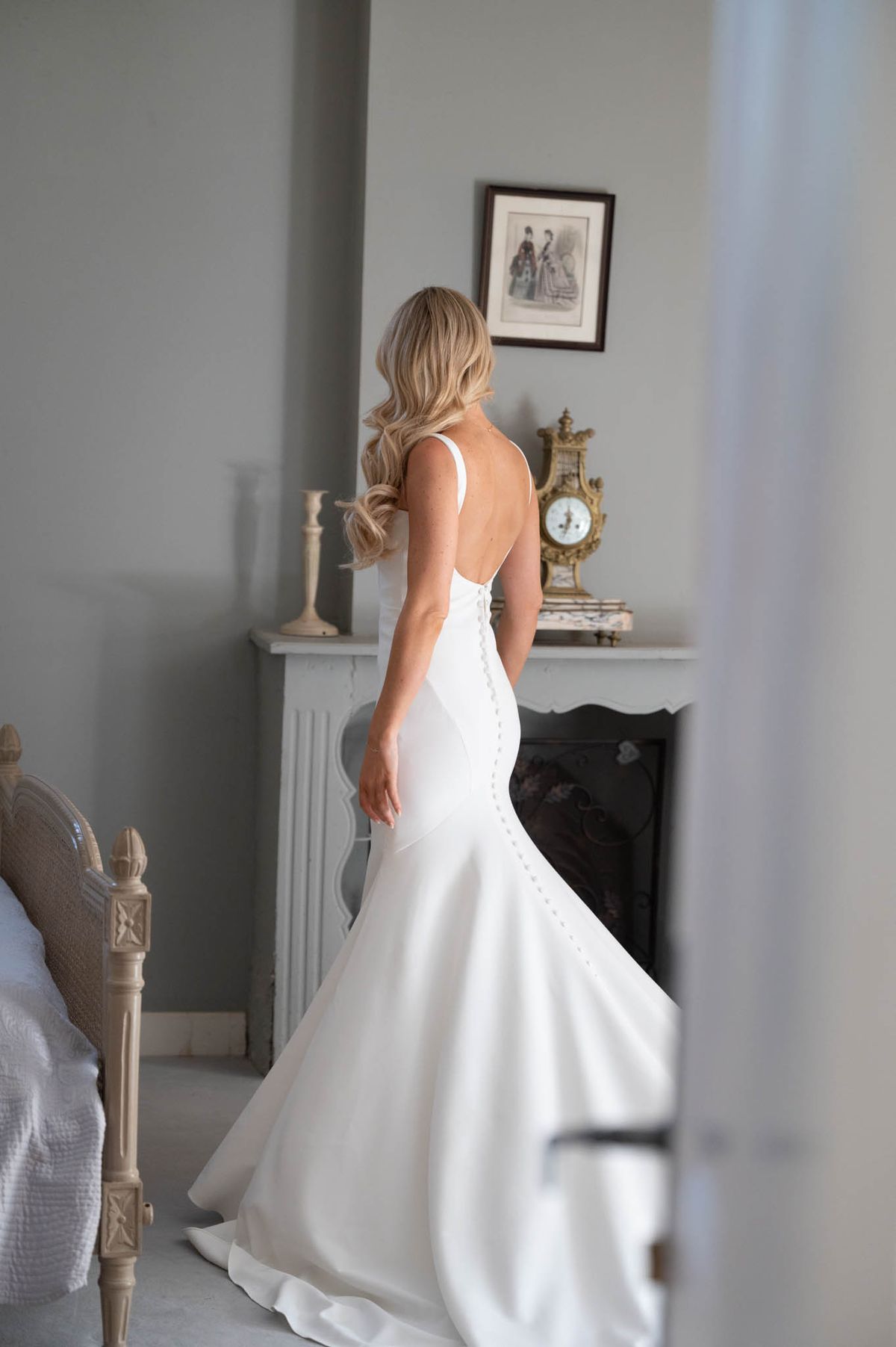 Bride seen from behind in low-back button-detail mermaid gown framed through a doorway in French bedroom