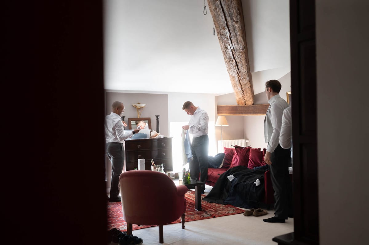 Three men are getting ready in a rustic indoor room, photographed through a doorway in a wide shot that frames the scene candidly. The room features exposed wooden ceiling beams, white walls, a dark wood chest of drawers with a small mirror, a red sofa with deep crimson cushions, a patterned red rug, and a floor lamp. All three men are in white dress shirts and grey trousers at various stages of dressing — one is holding a light blue item of clothing and looking down at it, one is arranging items on the chest of drawers, and one stands to the right with a dark waistcoat or cummerbund partially on. Dark suit jackets are laid across the sofa alongside other clothing items, and green bottles and a champagne bucket are visible on the coffee table, suggesting a pre-ceremony getting-ready moment.