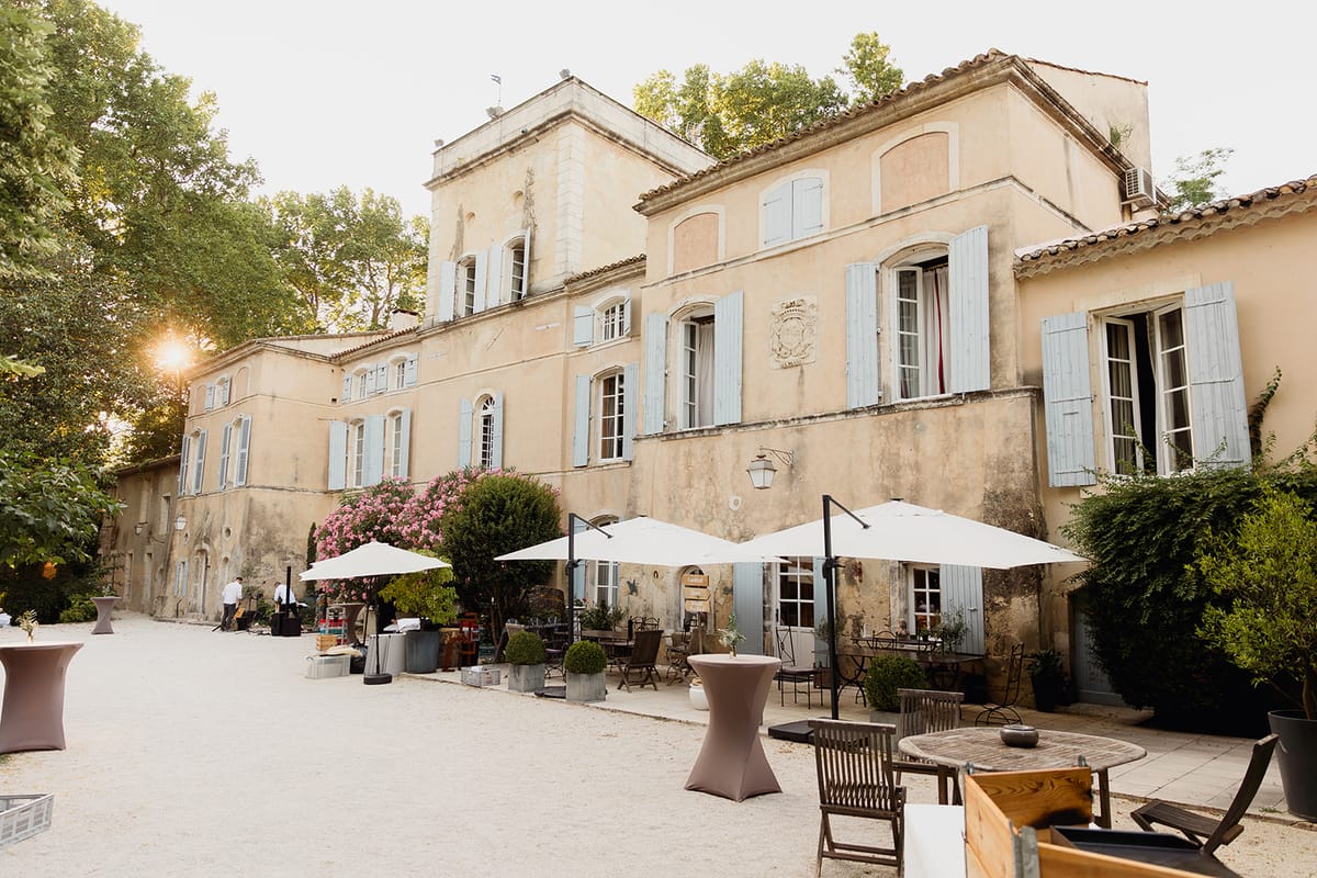 A cocktail hour is being set up or underway in the gravel courtyard of a French bastide or mas, a two-story ochre-rendered building with pale blue shutters and terracotta roof tiles. The outdoor space is furnished with cocktail-height poseur tables covered in taupe/mauve stretch linen, white market umbrellas on black cantilever stands, and wooden bistro chairs and tables arranged along the building's façade. Two staff members in white shirts are visible near a bar setup area on the left side of the courtyard, with what appears to be crates and bottles staged for service. Pink flowering shrubs grow along the base of the building, and clipped topiary balls in zinc planters line the entrance area. Warm golden-hour sunlight flares in from the left side of the frame. Wide-angle shot taken from courtyard level, emphasizing the full façade and event layout. Potential venue feature image.