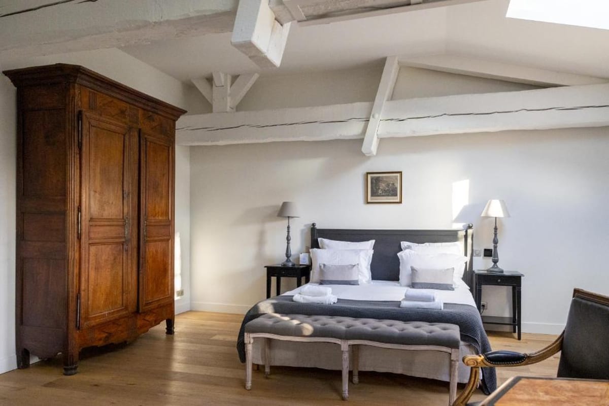 Interior shot of a guest bedroom, likely within a French château or manor house venue. The room features exposed white-painted timber ceiling beams, light oak hardwood flooring, and white walls. The bed has a dark charcoal iron frame dressed in white and grey linen with folded towels placed on top, flanked by two dark bedside tables with ivory lamp shades. A large antique French armoire in warm walnut wood stands to the left, and a grey tufted upholstered bench sits at the foot of the bed. A leather chair is partially visible in the foreground right. The styling mixes antique French provincial furniture with a clean, neutral grey and white palette. Wide interior shot. Potential venue feature image.