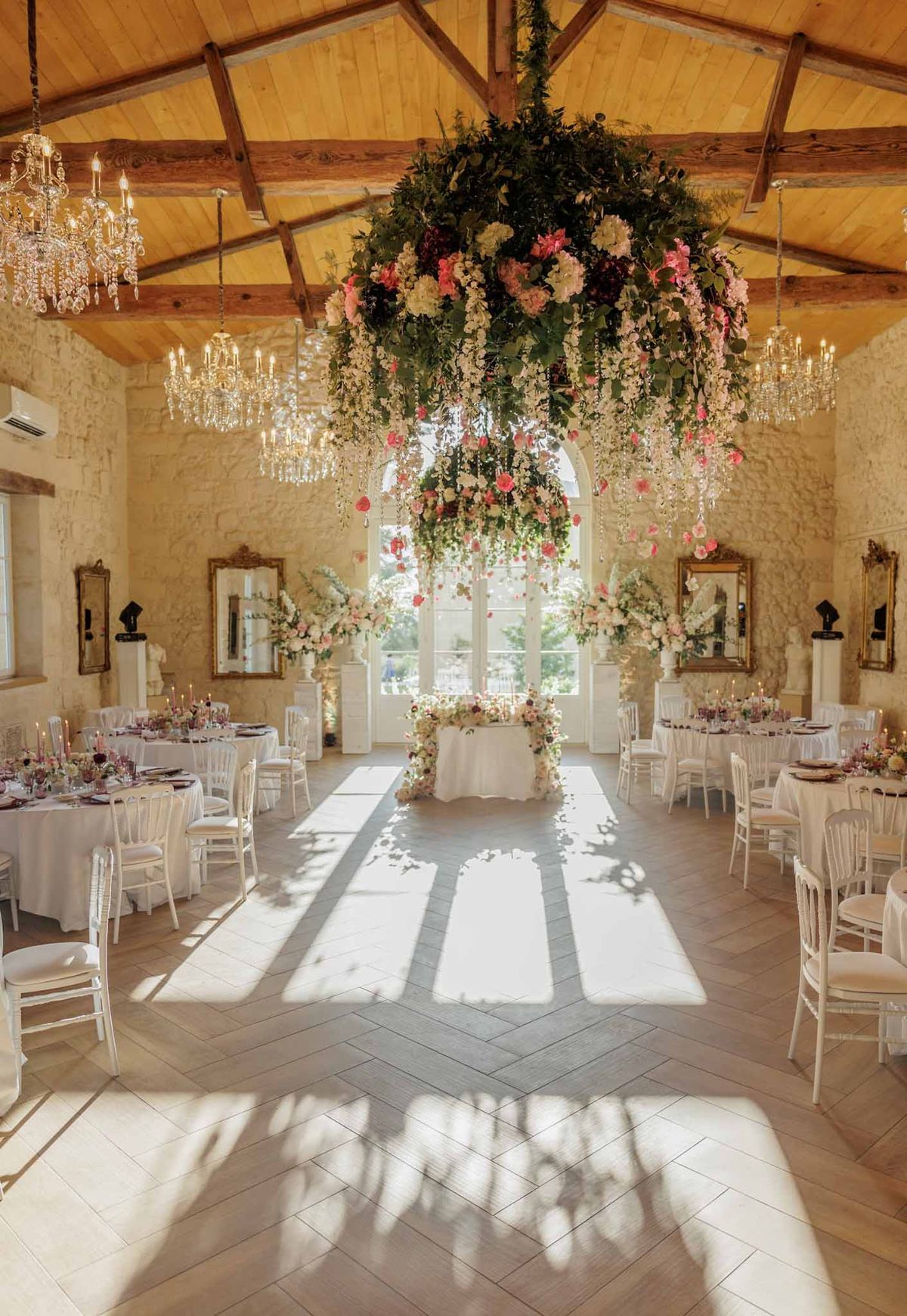 A wide-angle interior shot of a wedding reception room set up for a seated dinner, with no guests present. The space features exposed wooden beam vaulting, warm honey-toned tongue-and-groove ceiling panels, and limestone or cream stone walls hung with gilded ornate mirrors. The focal point is a large cascading floral installation suspended from the ceiling, composed of coral pink roses, ivory blooms, greenery, and trailing white wisteria-style florals with crystal drops. Crystal candelabra chandeliers flank the installation on either side. Round guest tables are dressed in white linen with white Napoleon/Chiavari chairs, set with candles and what appear to be deep burgundy-toned glassware and floral centerpieces in blush and jewel tones. A sweetheart or cake table at the far end is framed by tall white floral column arrangements and backed by a large arched French door letting in natural light. The herringbone-patterned light wood floor reflects strong afternoon sunlight streaming through the windows. The overall decor palette combines white, blush, coral, and burgundy in a classic French château ballroom style. Potential venue feature image.