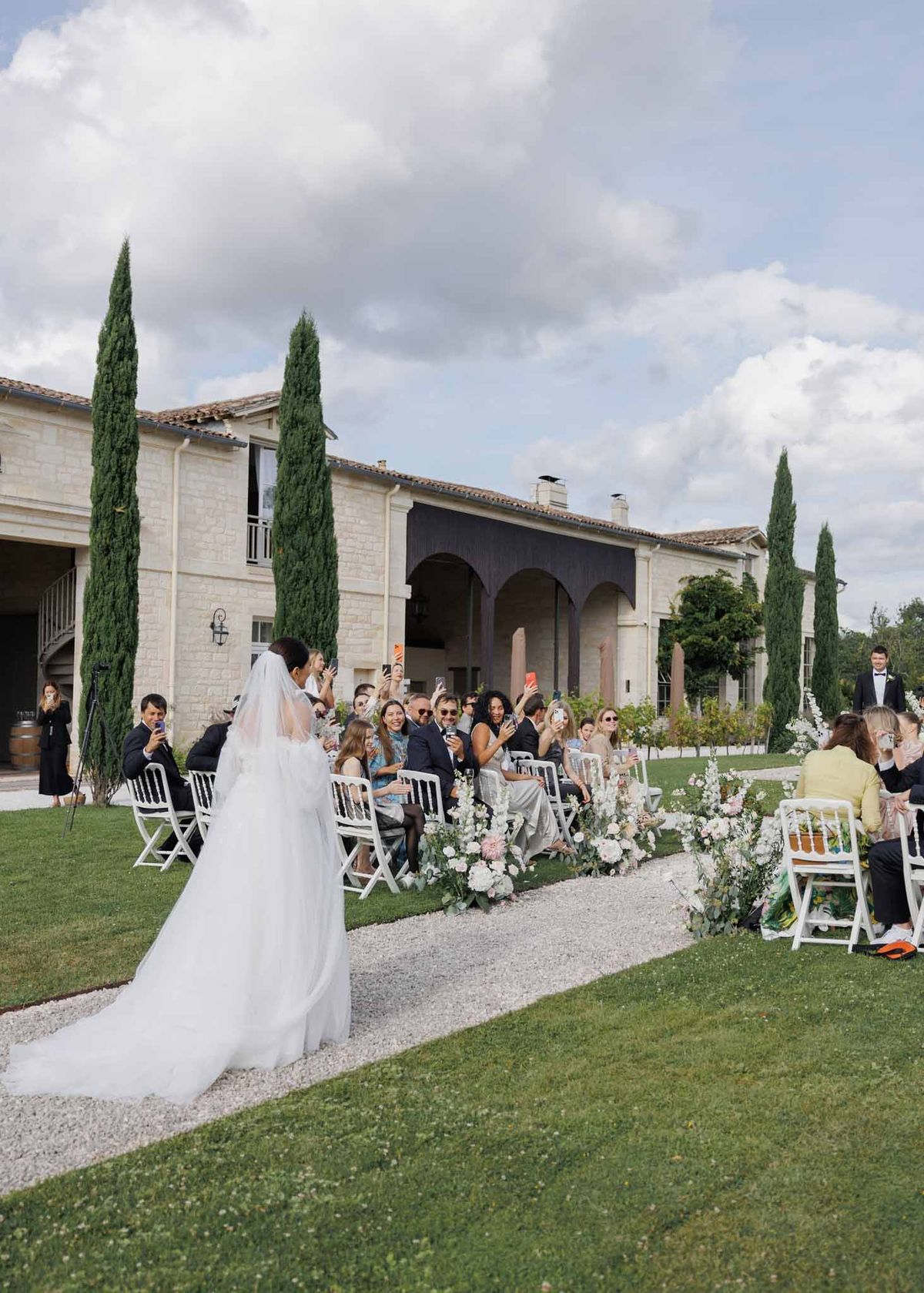 Bride walks down aisle with cathedral veil past blush dahlia arrangements toward stone manor with cypress