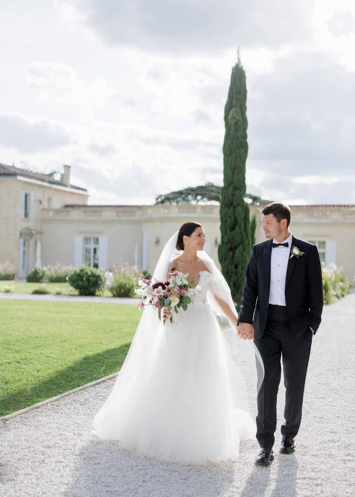 Bride in off-shoulder tulle ballgown and groom in black tuxedo walking hand-in-hand before French chateau facade