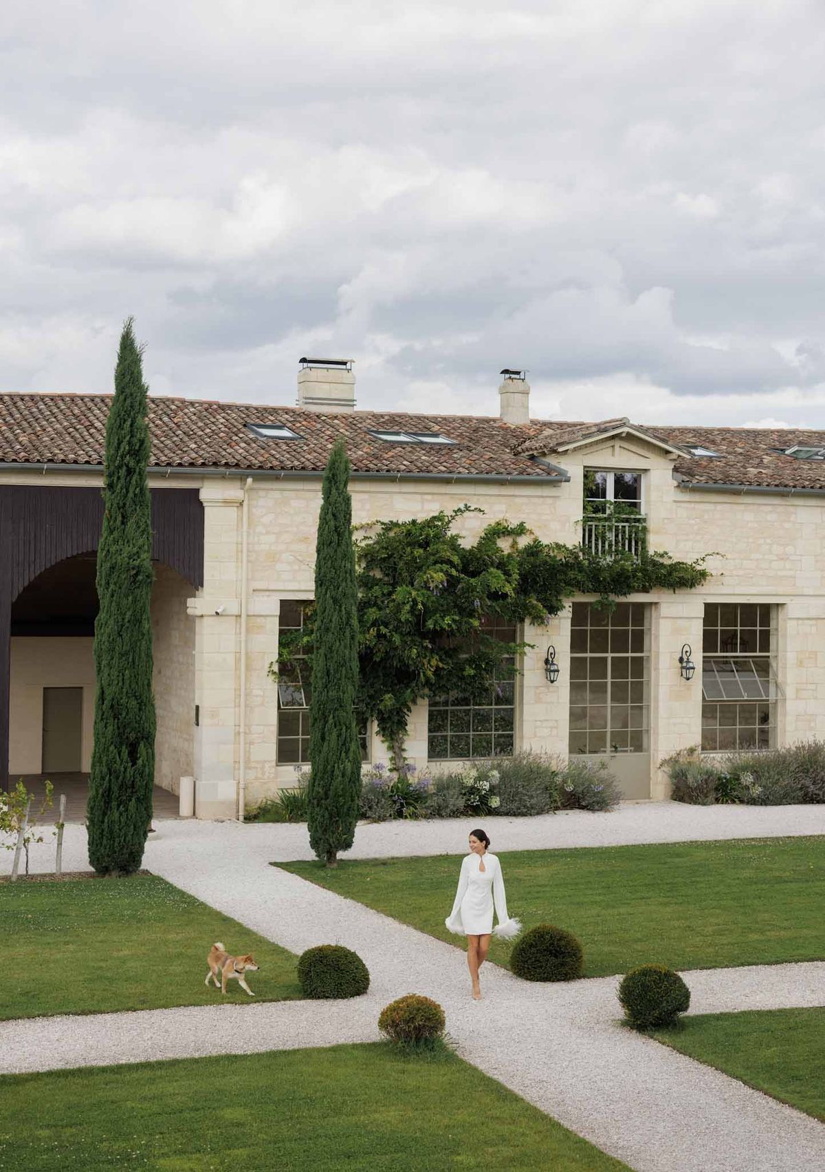 Bride in short white mini dress walking with Shiba Inu dog in formal gardens of French limestone estate