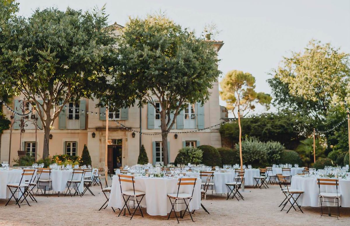 An outdoor wedding reception setup in the courtyard of a Provençal-style mansion featuring pale ochre stone walls and sage-green shutters. Approximately ten to twelve round tables are dressed in white floor-length linen tablecloths and surrounded by black metal bistro-style folding chairs with wooden slat seats and white cushions; tables are set with glassware and low greenery centerpieces. String lights are strung between wooden poles and across the facade of the building, and the sandy gravel courtyard is framed by trimmed topiary and mature trees. Wide shot taken at golden hour with no guests yet present. Potential venue feature image.
