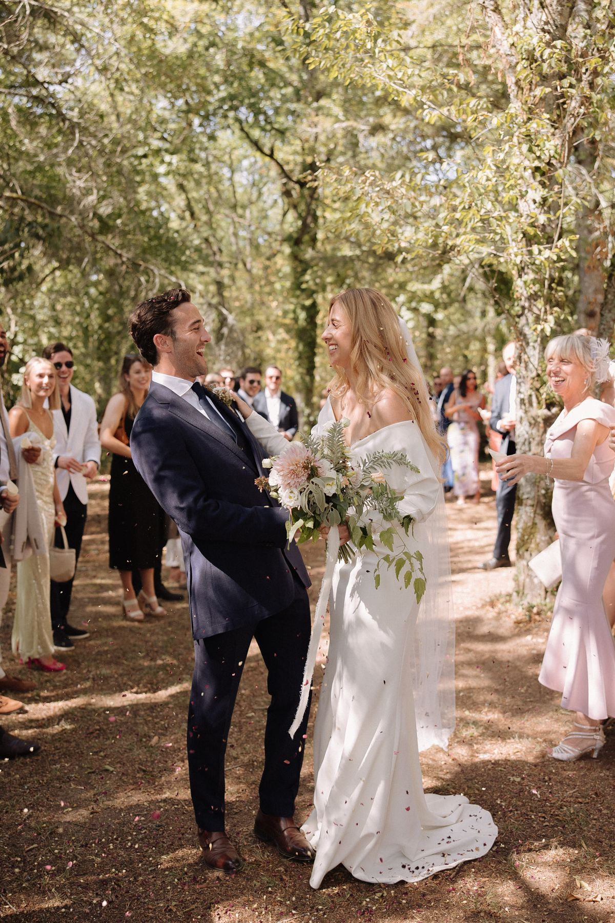 The couple is walking through a confetti send-off along a tree-lined outdoor aisle, laughing and facing each other just after their ceremony. The setting is a shaded woodland path with tall trees forming a natural canopy, lined on both sides by approximately 20–25 guests who are tossing small red and pink flower petal confetti. The bride wears an off-the-shoulder ivory satin slip-style gown with a train and a long sheer veil, and carries a loose, garden-style bouquet featuring blush dahlias, white blooms, and trailing fern and greenery. The groom wears a navy suit with a dark tie and brown leather Oxford shoes. Guests are dressed in a mix of casual summer attire, including a woman in a blush pink dress on the right. The overall styling is relaxed and natural with a boho-romantic feel. Wide portrait shot with natural dappled sunlight.