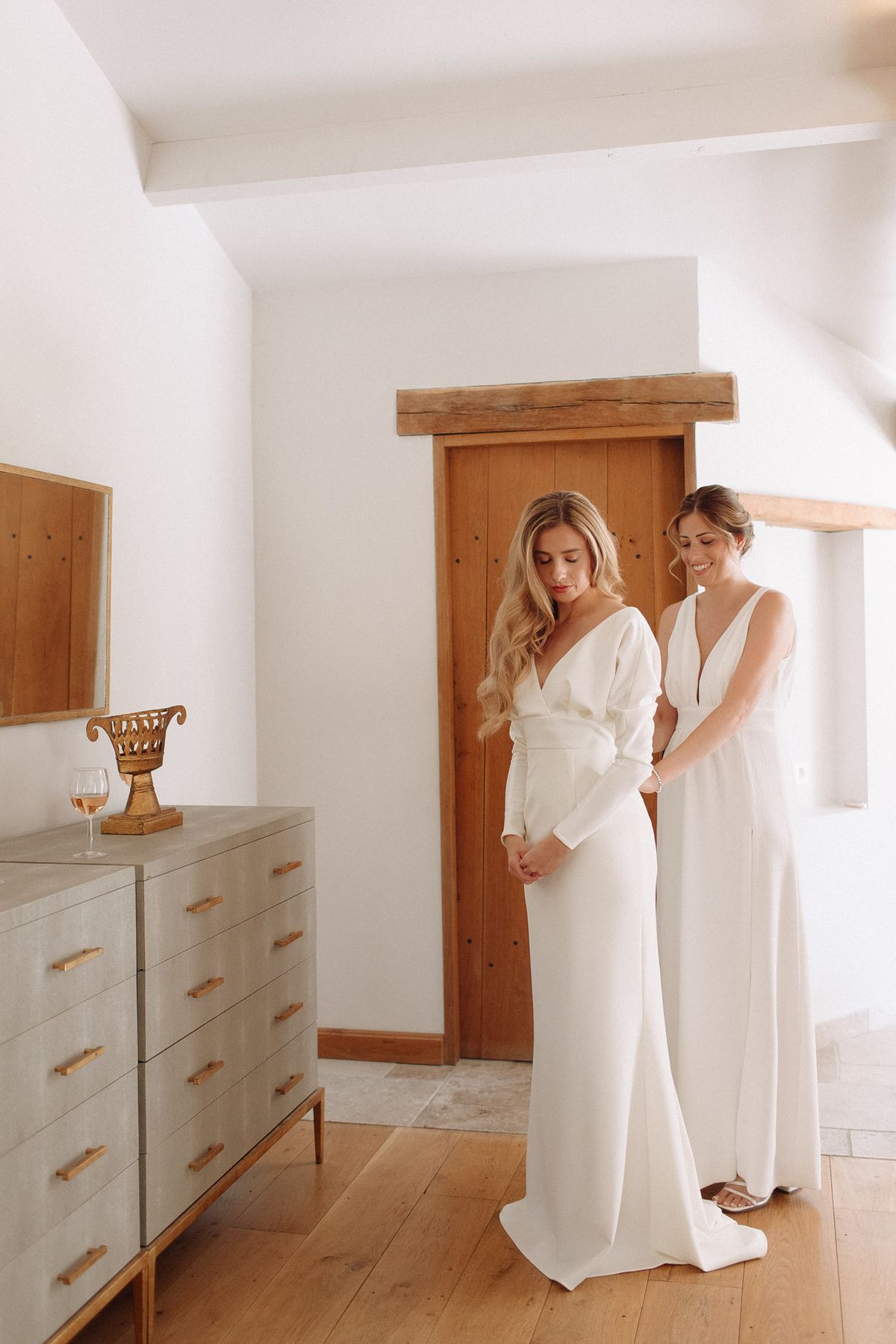 Companion fastening brides ivory crepe gown in bright rustic room with wooden beams chest of drawers and rose wine glass