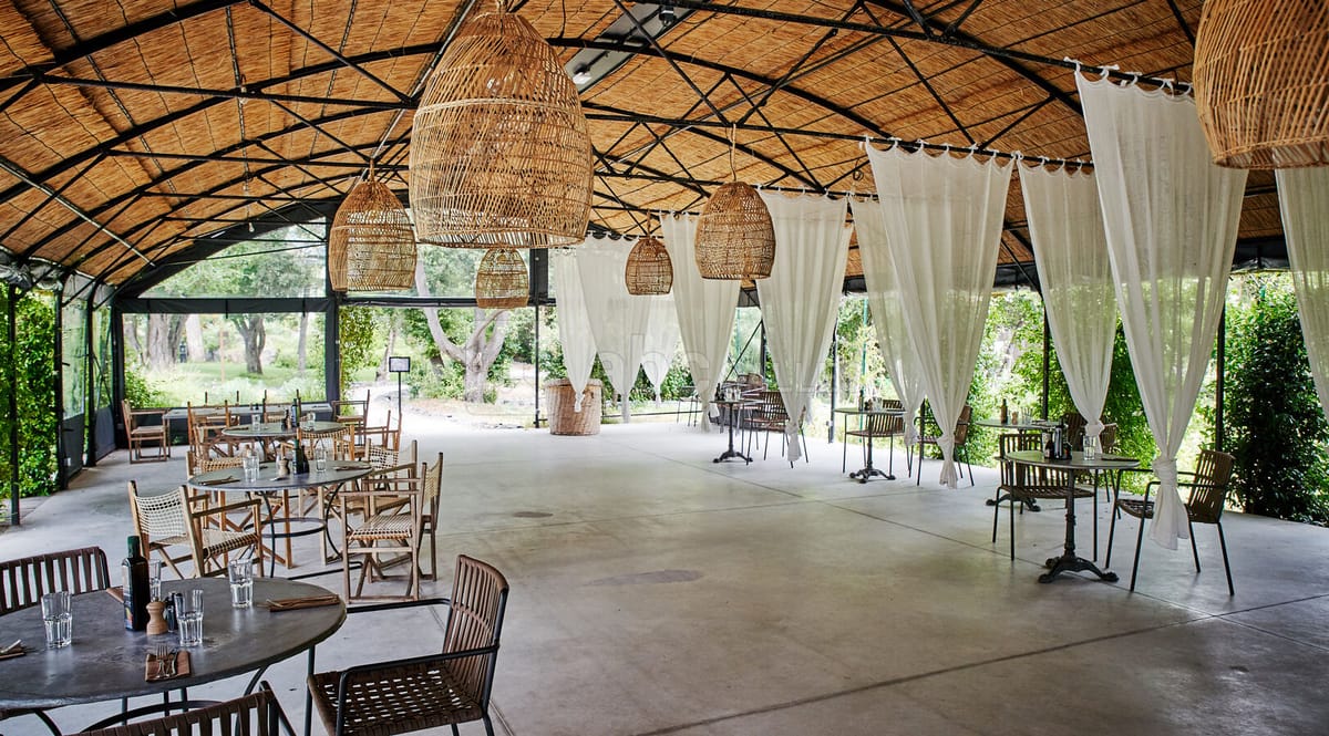 Thatched-roof open pavilion with wicker pendants, sheer curtains, and bistro chairs on polished concrete