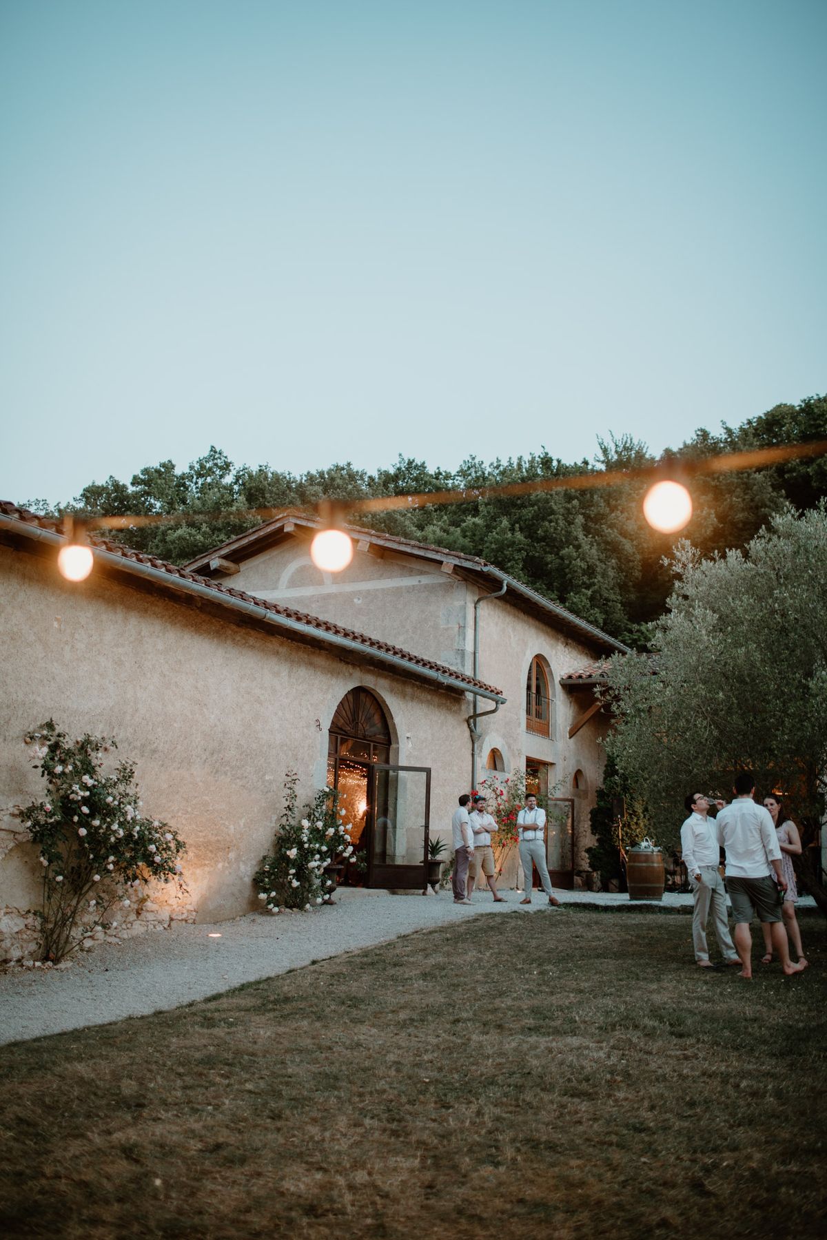 Guests mingling at dusk outside stone building with globe string lights and climbing roses