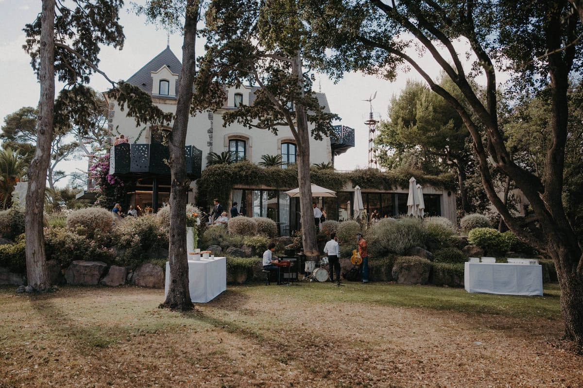 Guests mingling on manor terrace with live band on lawn cream parasols and turret-roofed white villa behind