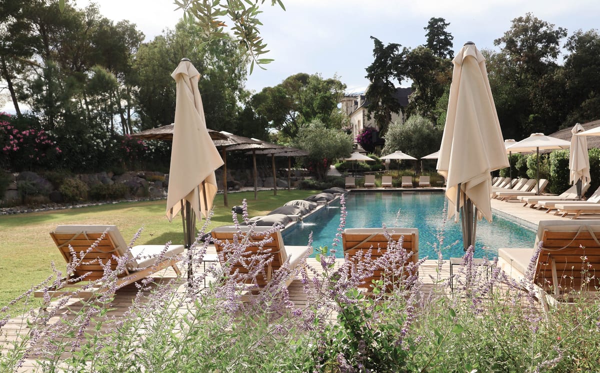 Outdoor venue grounds shot showing a long rectangular pool with blue water, surrounded by a wooden deck lined with teak sun loungers fitted with cream-colored cushions and closed cream canvas umbrellas. The pool area features dark grey oversized floor cushions at one end and a thatched pergola structure on the left side. The foreground includes purple salvia-style flowering plants. In the background, a classic stone manor house with a slate roof is partially visible through mature trees, with pink bougainvillea and oleander adding color to the mid-ground. The overall decor palette is neutral — cream, teak, and stone — with a relaxed Mediterranean styling. Wide shot taken from ground level at the garden edge. Potential venue feature image.