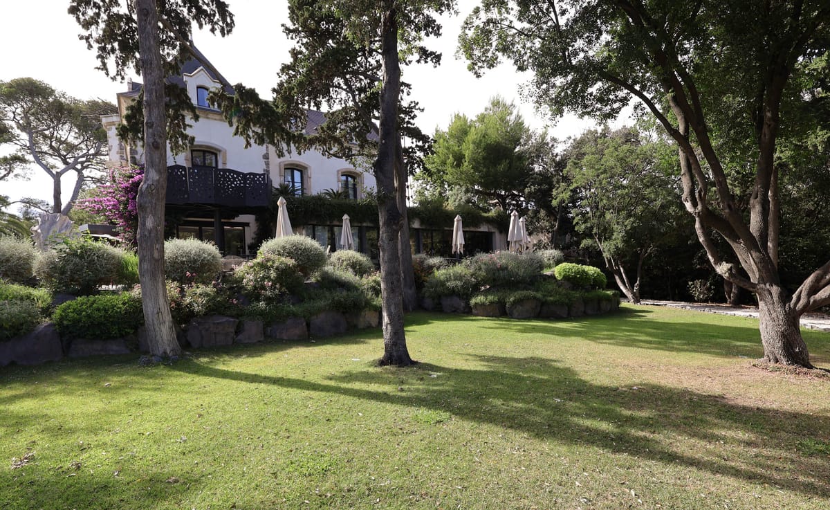 White-rendered villa with bougainvillea and cream parasols on tiered lawn with topiary and mature trees