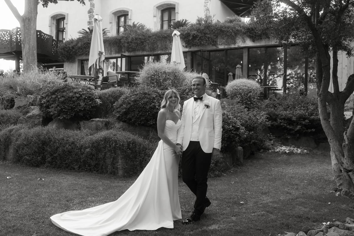 Bride in strapless gown and groom in white dinner jacket holding hands in chateau garden, black and white