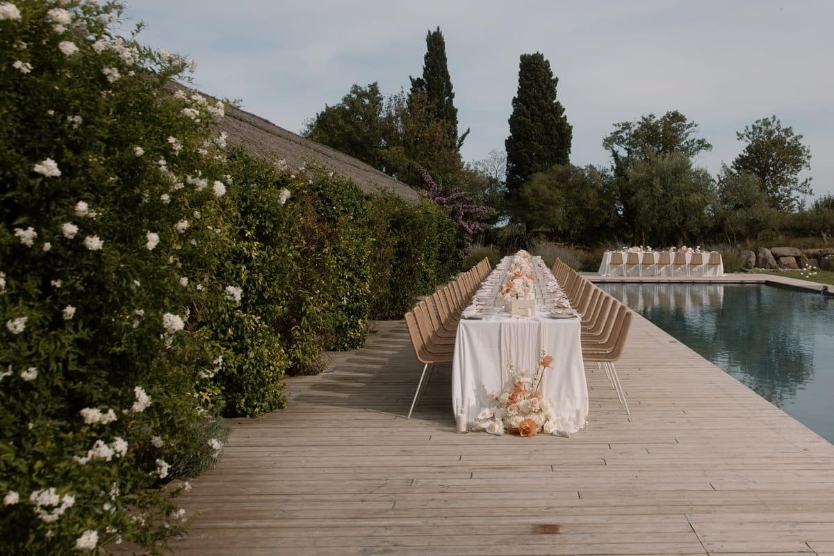 An outdoor wedding reception setup on a wooden deck running alongside a rectangular pool. A long feasting table covered in a white linen tablecloth is flanked by tan woven rattan-style chairs with white metal legs, set for approximately 20–24 guests. The table centerpiece features a runner of peach and ivory roses, with a cascading floral arrangement of peach garden roses, white ranunculus, and cream blooms pooling onto the deck at the near end. Place settings include glassware and white napkins visible along the table. In the background, a second table with white chairs — possibly ghost or acrylic chairs — is set further along the pool terrace. The decor palette is white, ivory, and peach, with a clean, modern aesthetic. A thatched-roof structure is visible to the left, bordered by a dense hedge with white flowering blooms. Wide shot taken from the end of the table looking down its length. Potential venue feature image.