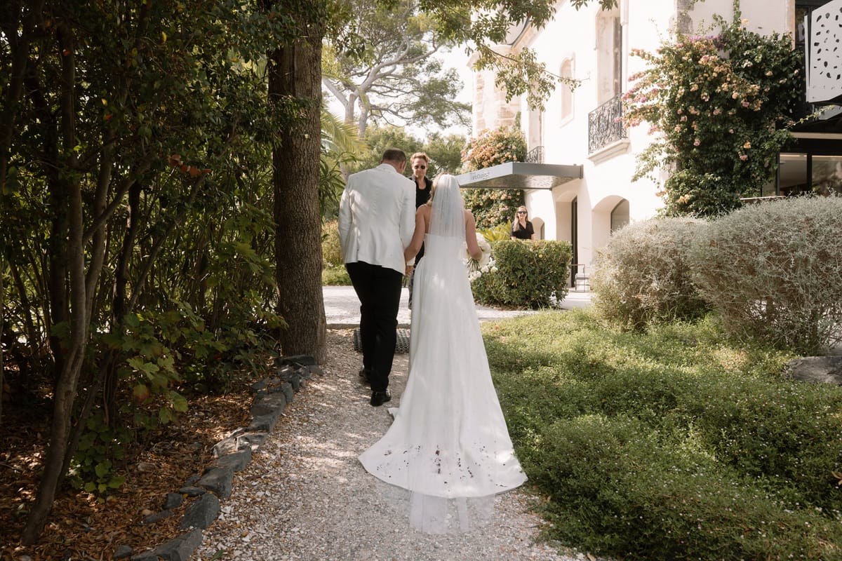 Bride and groom walking toward white villa entrance with pink climbing vines from behind