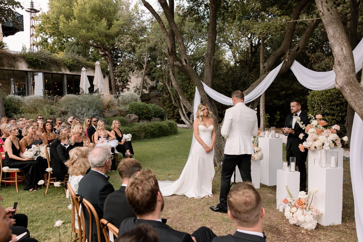 An outdoor wedding ceremony taking place in a garden setting, with approximately 40–50 guests seated in rows on wooden chairs facing the altar. The bride wears a white strapless fitted gown with a long train and stands facing the groom, who is dressed in a white blazer with black trousers; an officiant in a black tuxedo stands to their right reading from a booklet. The altar area features white fabric draped between mature trees, white geometric plinths at varying heights holding pillar candles, and arrangements of peach, coral, and ivory roses and ranunculus. Bridesmaids seated among guests wear black spaghetti-strap dresses and hold small ivory floral bouquets, while most male guests are dressed in black suits or tuxedos. The ceremony backdrop includes a contemporary building with large glass windows and warm interior lighting visible in the background. Wide shot taken from behind and to the side of the seated guests, capturing the full ceremony scene.