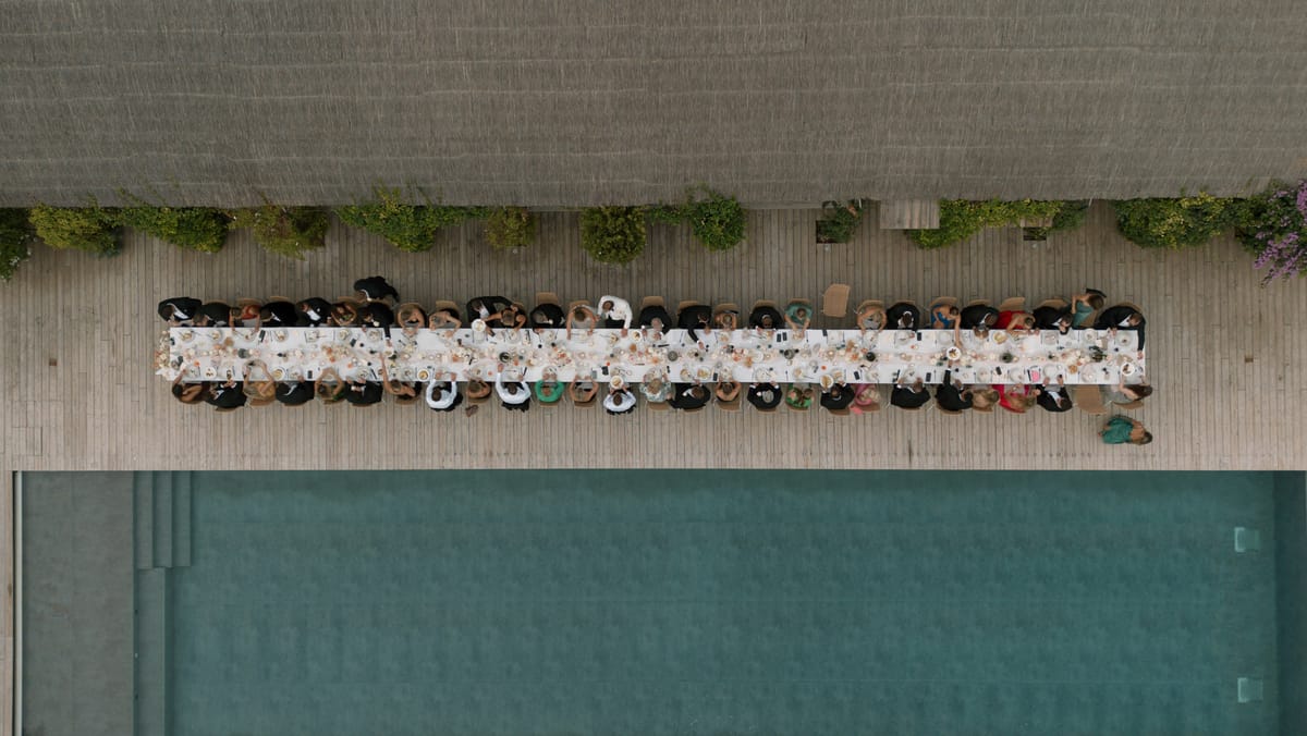 Aerial view of wedding reception dinner along a long table on wooden deck beside a swimming pool