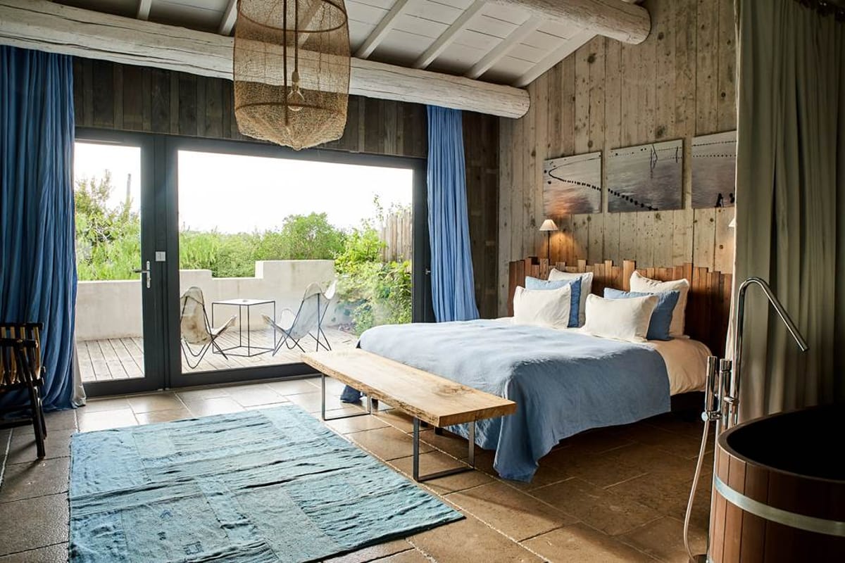 Rustic-modern bedroom with blue linens, barrel bathtub, and deck opening to countryside