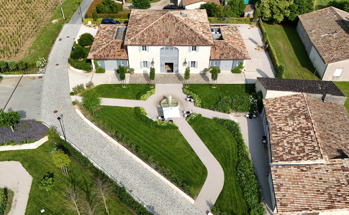 Aerial drone shot of a French country estate or château property, showing a white rendered main building with terracotta tile roofing, grey shutters, and a large arched entrance. The formal forecourt features a central stone fountain surrounded by manicured lawn panels, clipped topiary trees lining the entrance path, and white flowering borders. A white parasol with dark bistro chairs and tables is set up near the fountain, suggesting a cocktail hour or outdoor seating arrangement. The property is flanked by additional outbuildings with matching terracotta roofs, and vineyards are visible to the upper left of the frame. Potential venue feature image.