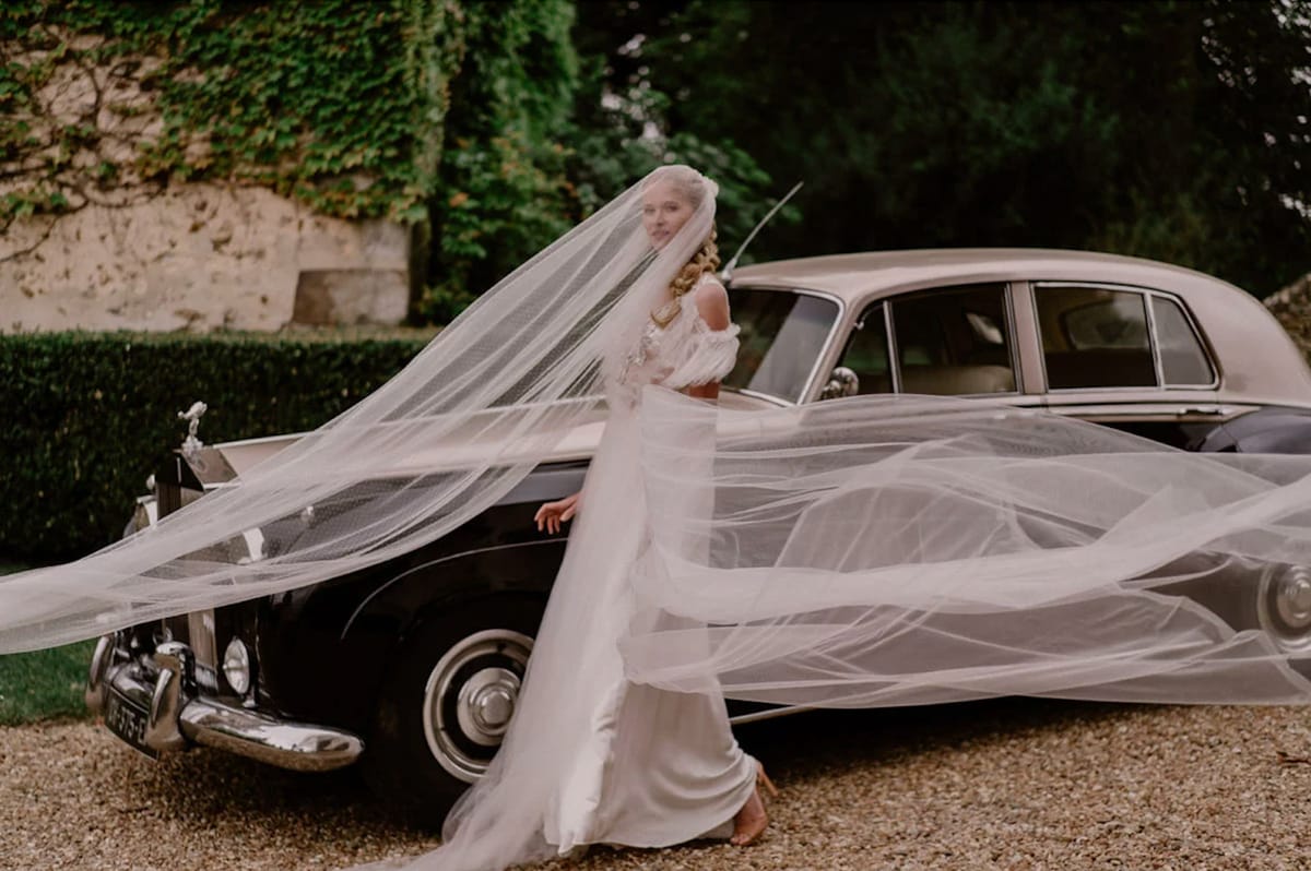 Bride beside vintage navy Rolls-Royce with cathedral veil billowing in wind on chateau gravel driveway