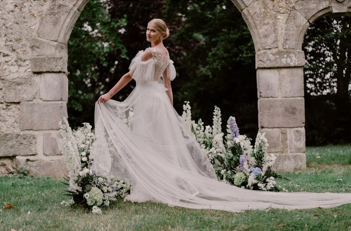 Bride in off-shoulder tulle ballgown with cathedral train poses before stone archway with blue and white floral installation