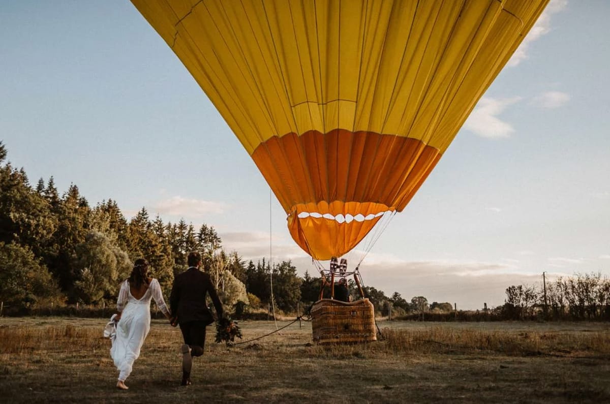 Couple walking toward amber hot air balloon in rural field at golden hour, bride barefoot with foliage bouquet