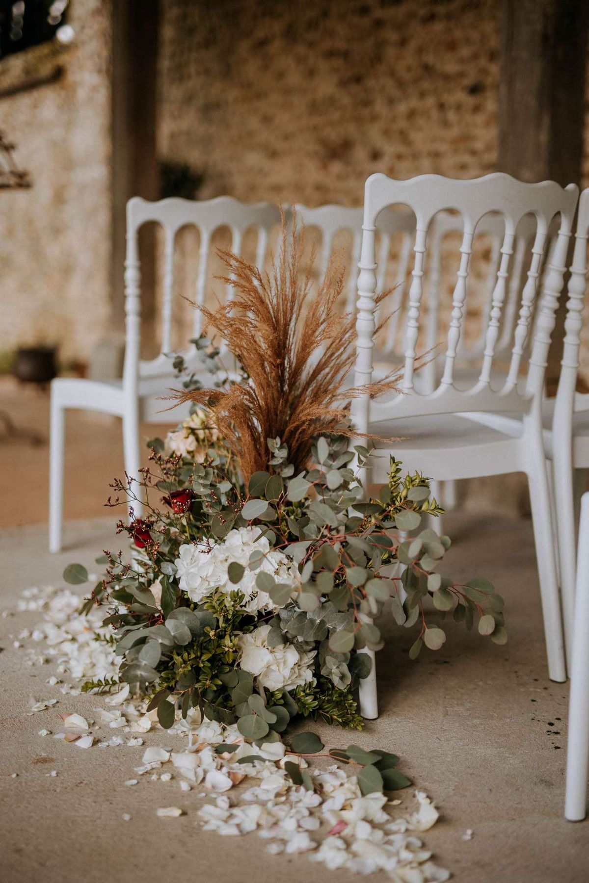A close-up detail shot of a ceremony aisle floral arrangement placed on the floor beside white Napoleon chairs at an indoor-outdoor venue with exposed stone walls. The arrangement combines white hydrangeas, deep red roses, eucalyptus branches, small-leafed green foliage, dried pampas grass, and burgundy berry stems, creating a boho-rustic aesthetic. Loose white flower petals are scattered across the concrete floor around the base of the arrangement, extending along the aisle. The color palette mixes ivory, deep red, sage green, and warm caramel tones from the dried grasses.