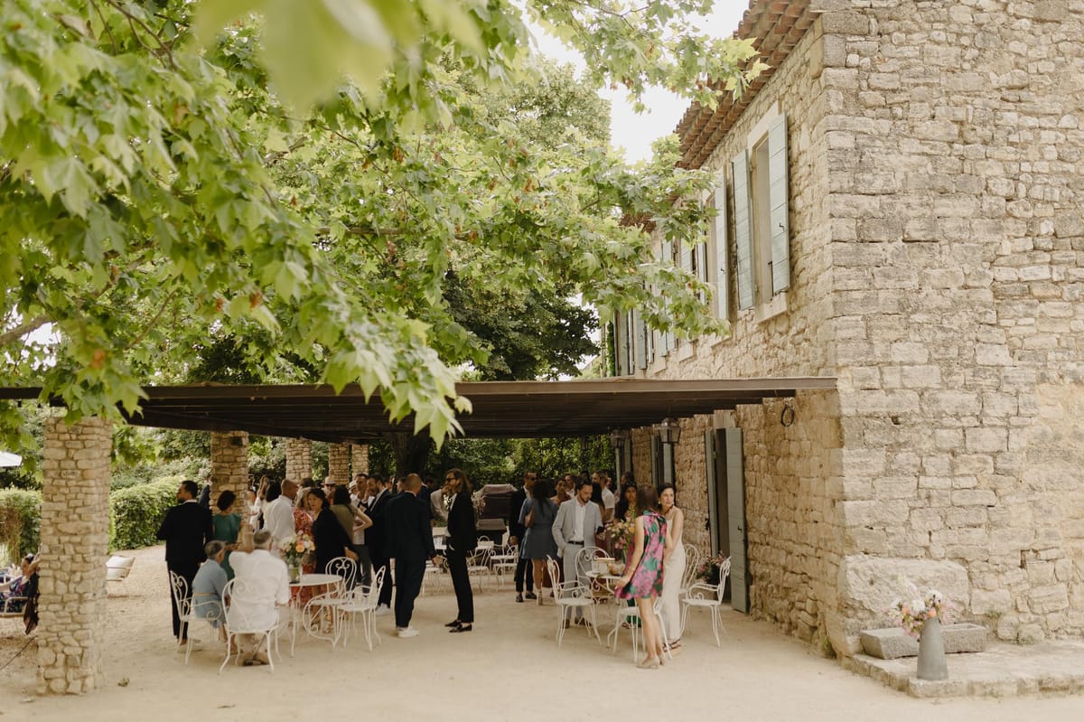 A cocktail hour is taking place outdoors at a French stone-building venue, with approximately 25–30 guests mingling and seated beneath a flat-roofed pergola structure attached to the main building. The setting is a courtyard alongside a two-storey limestone mas or bastide-style property with pale blue shutters. Guests are dressed in smart-casual summer attire, including a woman in a bright pink and green floral dress in the foreground and others in dark suits and light linens. White wrought-iron bistro chairs and small round tables are arranged under the covered area. A metal pitcher holding a small arrangement of peach and pink flowers sits on a stone step near the building's entrance, with a matching floral arrangement visible on one of the tables. The overall styling is relaxed and classic, with a warm, informal atmosphere. Wide shot taken from a slight distance with tree branches framing the top of the frame. Potential venue feature image.