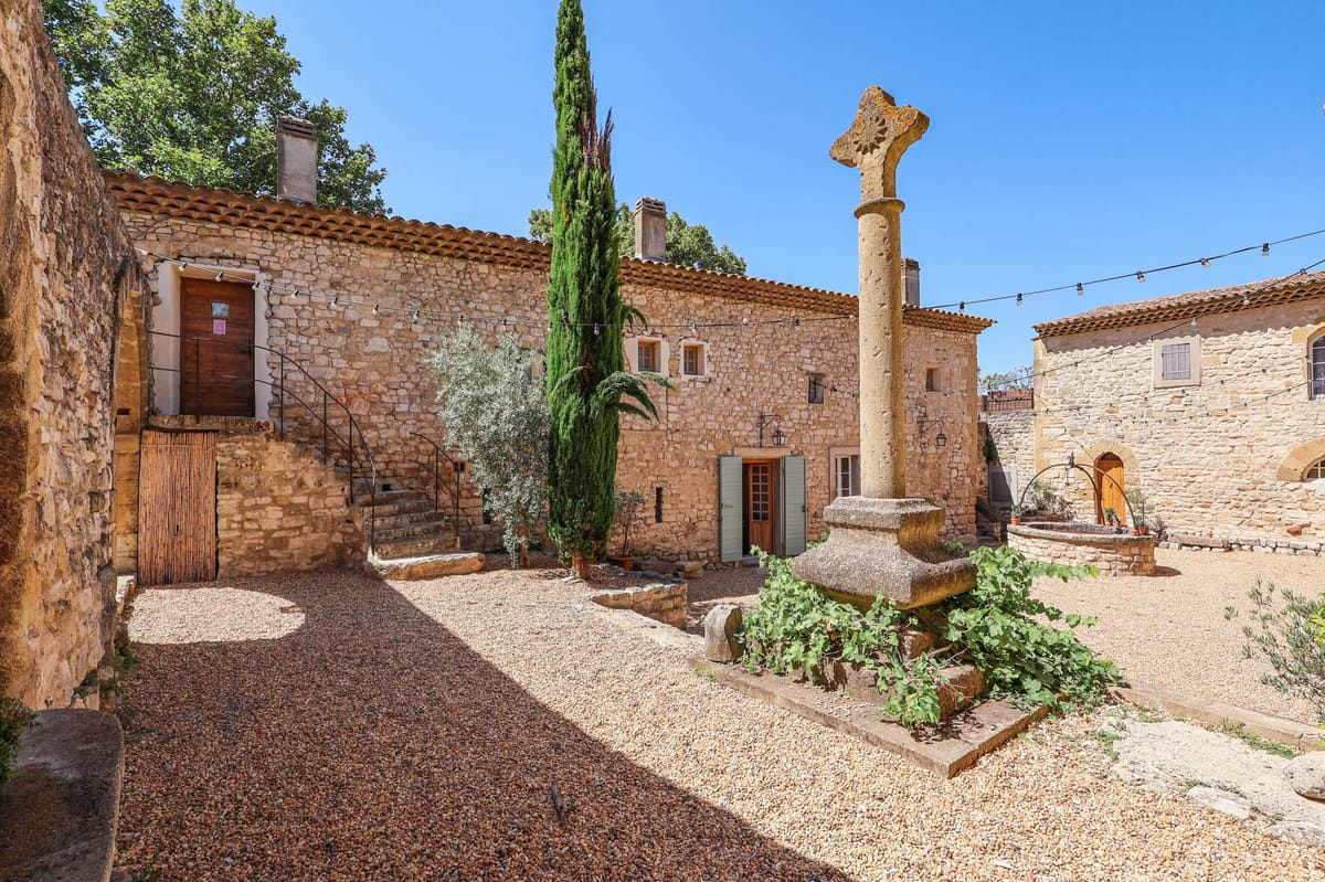 A wide-angle exterior shot of a historic Provençal stone property featuring a gravel courtyard flanked by multiple honey-colored limestone buildings with terracotta-tiled roofs and sage green shutters. A carved stone cross monument on a column pedestal stands prominently in the foreground, surrounded by climbing ivy. Festoon lights are strung between the buildings, indicating event use. A tall cypress tree and an olive tree are planted within the courtyard space, and a stone archway with a wooden door is visible on the right side of the frame. No people are present. Potential venue feature image.