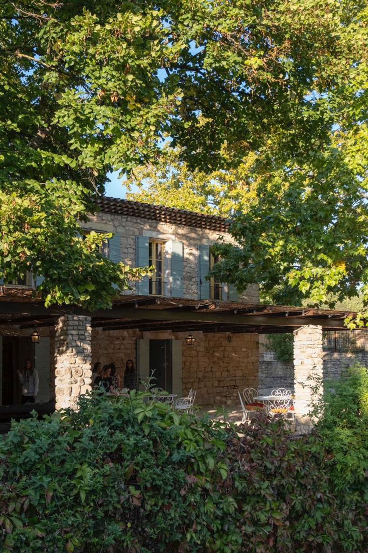 Two-story stone bastide with terrace portico, wrought-iron garden chairs, and garden foliage