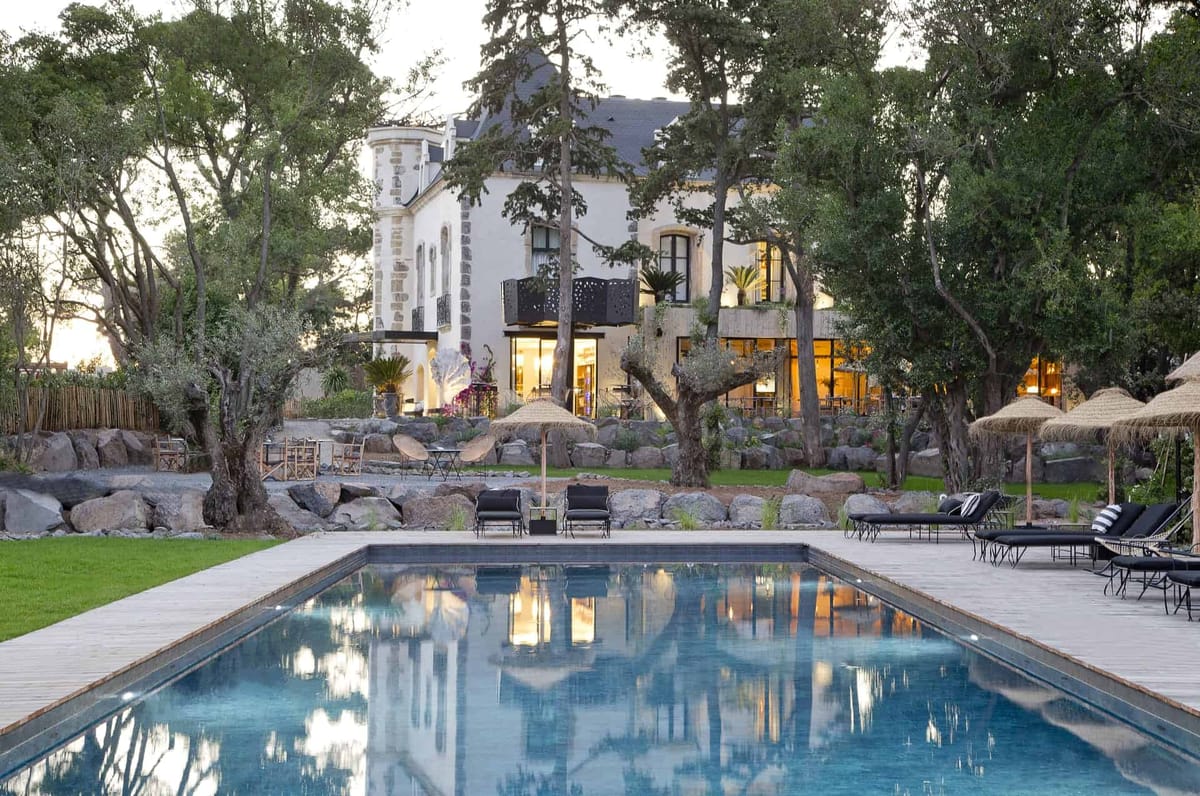 White manor with turret, pool, straw parasols, and olive tree terraced garden photographed at dusk