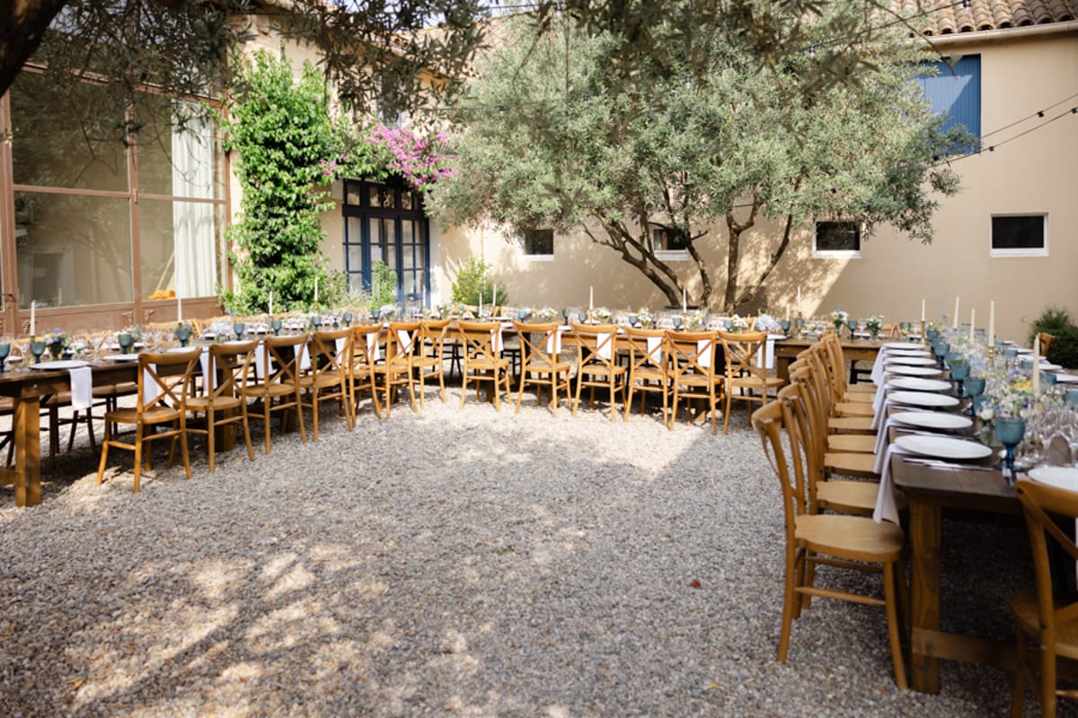 An outdoor wedding reception setup in a gravel courtyard surrounded by the warm ochre-toned walls of a Provençal mas or farmhouse. Three long wooden farm tables are arranged in a U-shape, each lined with natural wood cross-back chairs. The tables are dressed with white linen runners, white plates, clear glassware, dusty blue goblets, tall white taper candles, and low floral centerpieces in muted tones. No guests are present, as the image captures the fully set tables before the reception begins. The decor palette is rustic-natural with a blue and white accent scheme. The shot is taken from a wide, slightly elevated angle showing the full table arrangement. Potential venue feature image.