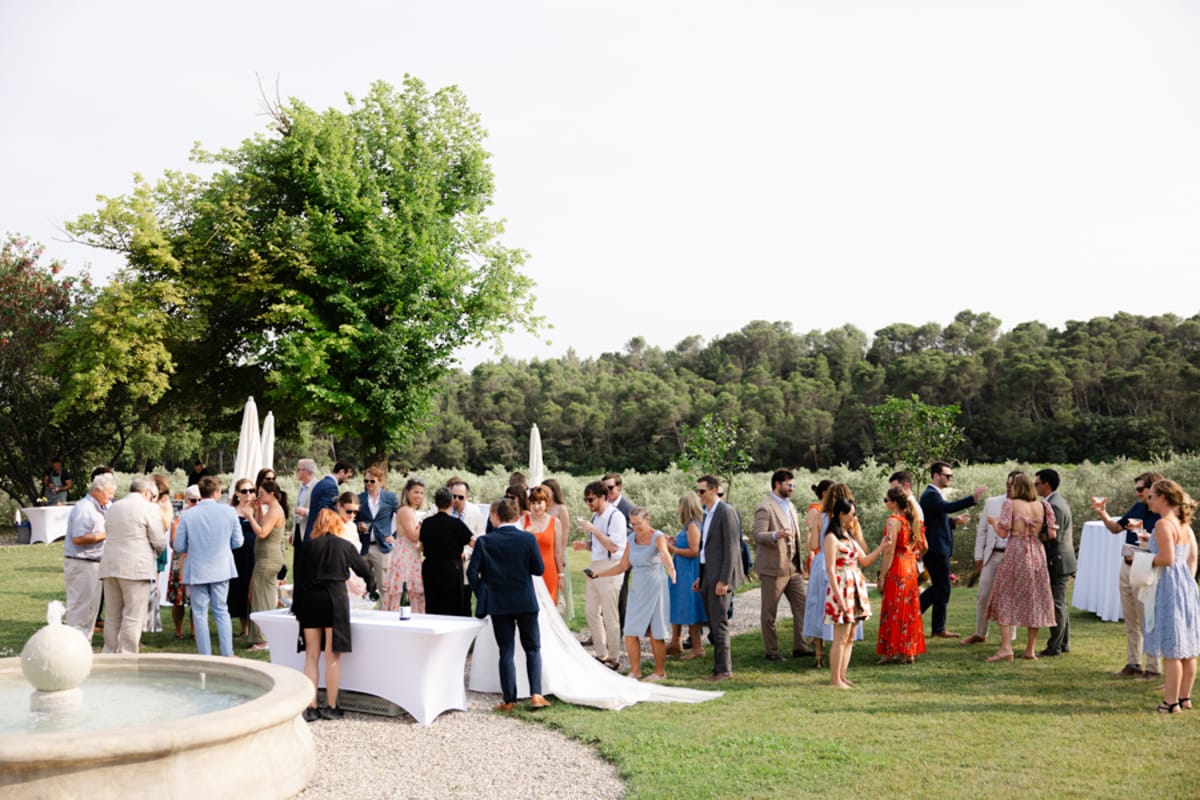 An outdoor cocktail hour taking place on a lawn area beside a stone fountain, with approximately 40–50 guests mingling in small groups. The setting appears to be a Provençal property surrounded by olive trees and woodland, with white cocktail tables dressed in floor-length white linen cloths and closed white market umbrellas visible in the background. The bride, wearing a white gown with a long train, is visible near the center of the frame. Guests are dressed in smart-casual summer attire including a bright orange midi dress, a red floral maxi dress, a pale blue dress, and various linen suits in tan and navy. The shot is a wide environmental frame capturing the full scene from a slight distance.