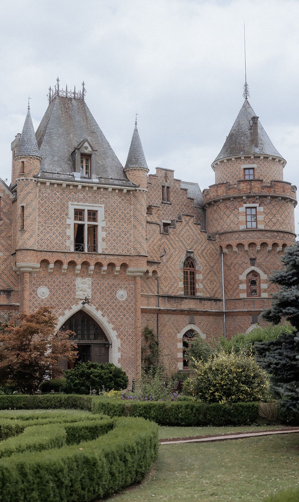Exterior wide shot of a French château featuring warm terracotta and red brick construction with decorative diamond-pattern brickwork, multiple conical-roofed towers with grey slate finials, Gothic pointed archway entrance, and arched window details with white stone surrounds. The foreground includes formally clipped boxwood hedges arranged in geometric shapes, with ornamental shrubs and a blue-grey conifer to the right. No people are visible in the frame. The image is taken in natural overcast light. Potential venue feature image.