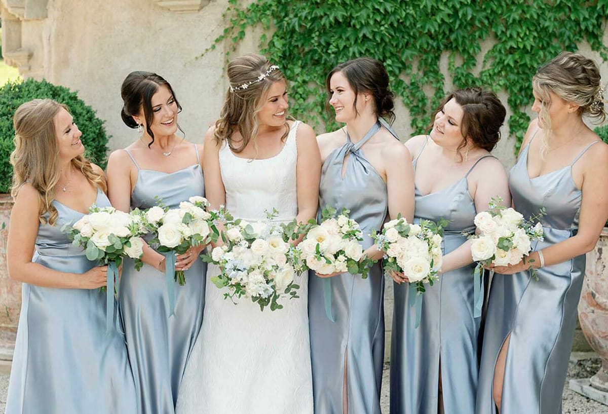 A bridal party portrait taken outdoors against an ivy-covered stone wall, likely at a French château or historic estate. The bride stands at the center wearing a sleeveless, fitted ivory lace gown with a delicate floral hair accessory, surrounded by five bridesmaids in floor-length dusty blue satin dresses in varied neckline styles including spaghetti straps and a halter tie. Each bridesmaid holds a round bouquet of ivory garden roses, soft blush roses, eucalyptus, and small blue accent flowers, while the bride's bouquet is slightly larger with trailing sage green ribbon. The group is laughing and looking at one another in a candid, relaxed moment, captured in a medium group portrait with a classic and polished styling aesthetic.
