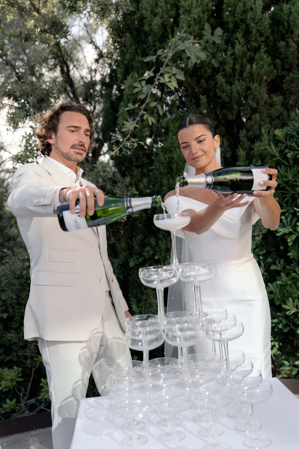 The bride and groom are pouring champagne into a coupe glass tower during what appears to be a cocktail hour or reception moment, set outdoors against a backdrop of dense green foliage. The groom wears an ivory suit with an open-collar white shirt, and the bride wears a fitted off-the-shoulder white satin gown with a long veil, her dark hair pulled back sleekly. Both are holding green champagne bottles — consistent with a Champagne house label — and pouring simultaneously into the top tier of a pyramid of clear coupe glasses arranged on a white-clothed table. The shot is a medium portrait framing both figures from approximately the waist up, with the glass tower prominently featured in the foreground.