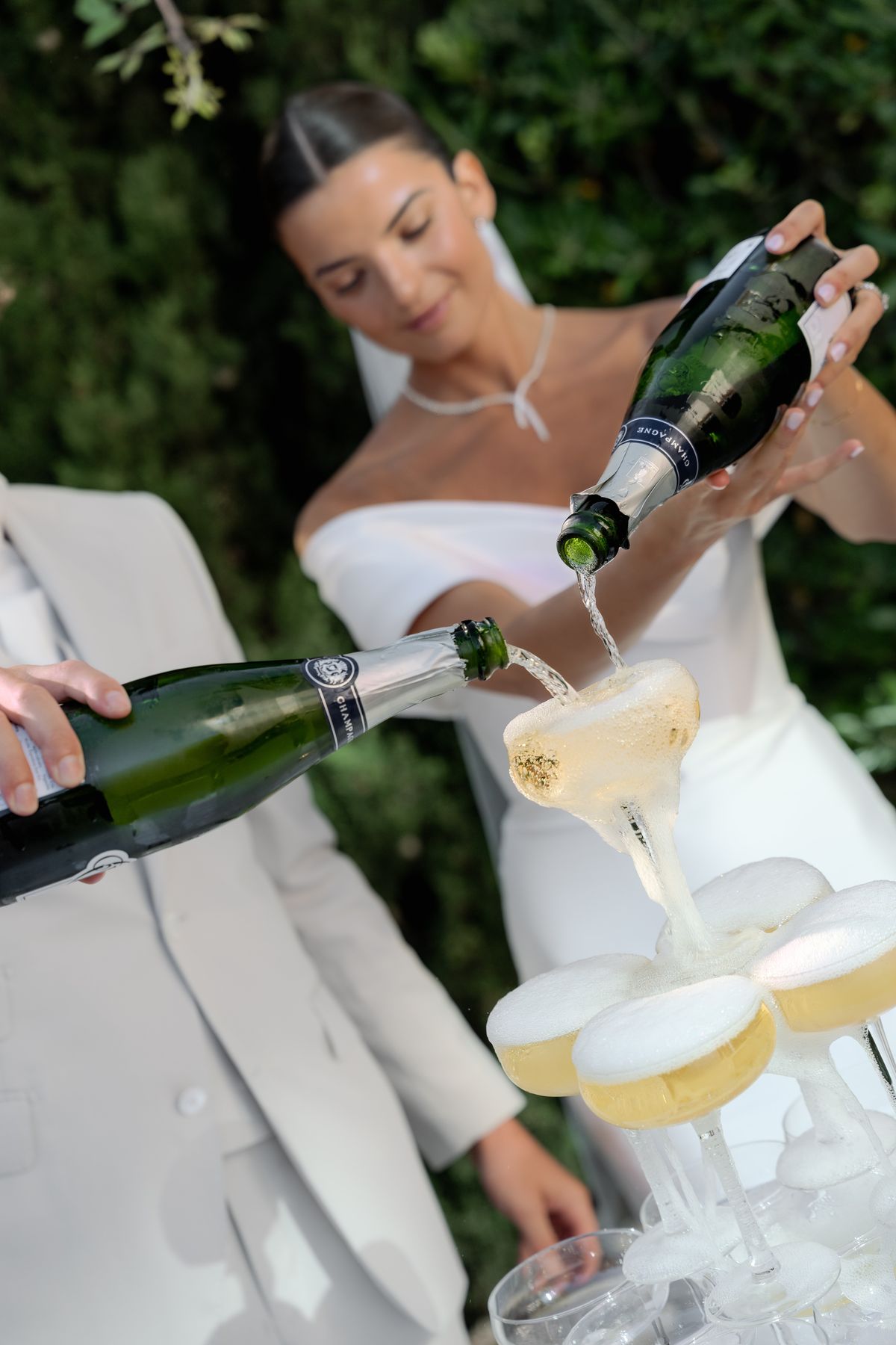 The bride and groom are pouring champagne simultaneously into the top tier of a champagne tower during what appears to be a cocktail hour or reception moment outdoors. The bride wears a white strapless fitted dress with a delicate white necklace and has her dark hair pulled back sleekly; the groom is in a light ivory or off-white suit. Both are holding dark green champagne bottles labeled 'Champagne,' and the liquid is visibly overflowing and cascading down into the coupe glasses below. The tower consists of multiple tiers of wide champagne coupe glasses filled with golden, frothy champagne. The shot is a close-up portrait-style image with the action in sharp focus in the foreground and the bride softly blurred in the background.