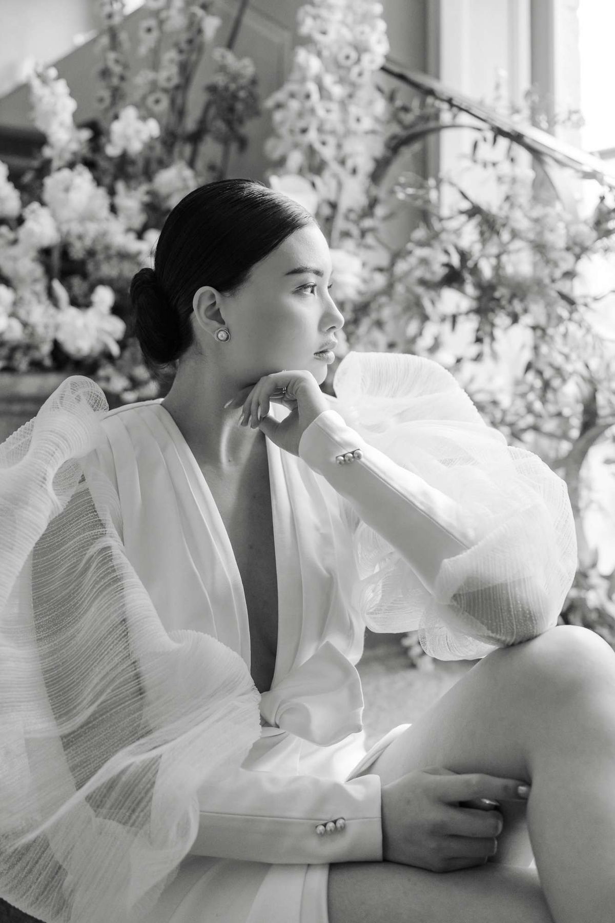 Black and white portrait of bride in structured white blazer seated in profile before indoor white floral installation