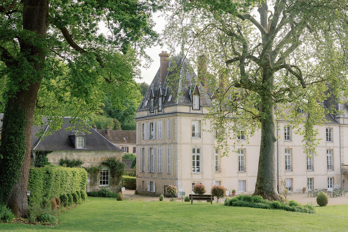A wide exterior shot of a French château complex featuring a cream-colored stone main building with a distinctive pointed slate-roofed tower, tall shuttered windows, and a multi-story facade. The grounds include a manicured lawn in the foreground, a gravel courtyard area with a wooden bench, trimmed boxwood hedges, and outdoor seating visible to the right side of the building. A secondary stone outbuilding with a lower roofline sits to the left. No people are present in the image. The overall architectural style is classic French country estate. Potential venue feature image.