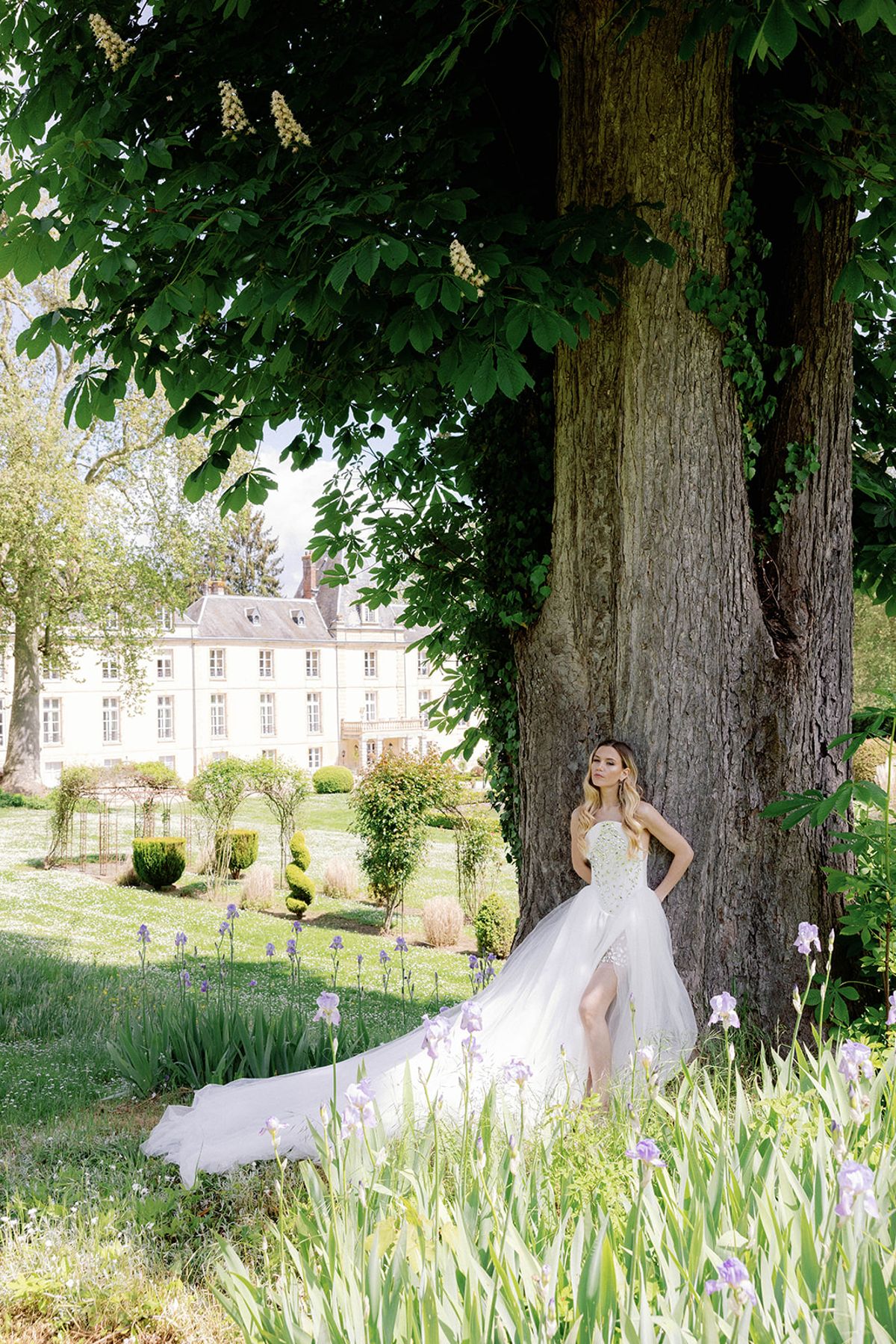 Bride in strapless lace ballgown leaning against tree trunk with iris blooms in chateau formal garden