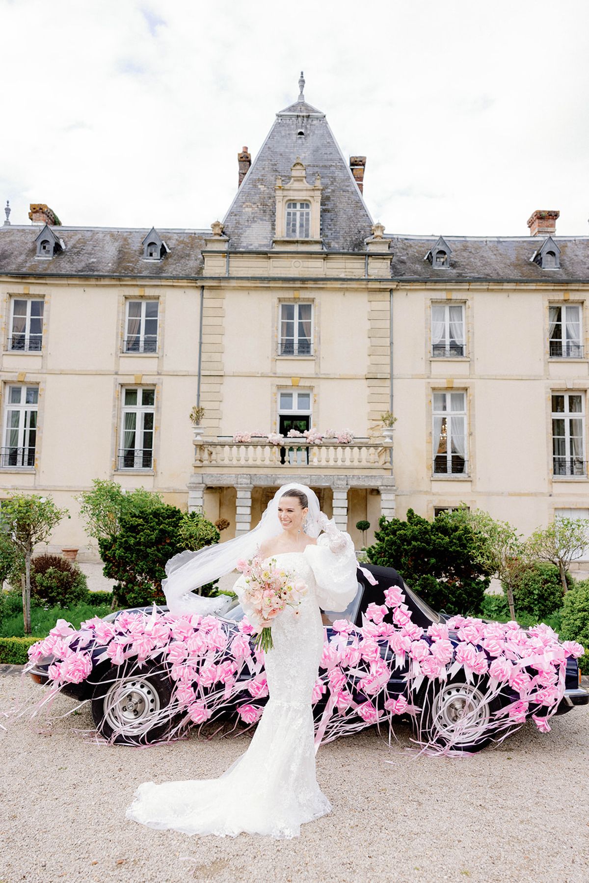 Bride in lace mermaid gown with cathedral veil before chateau and rose-decorated vintage convertible