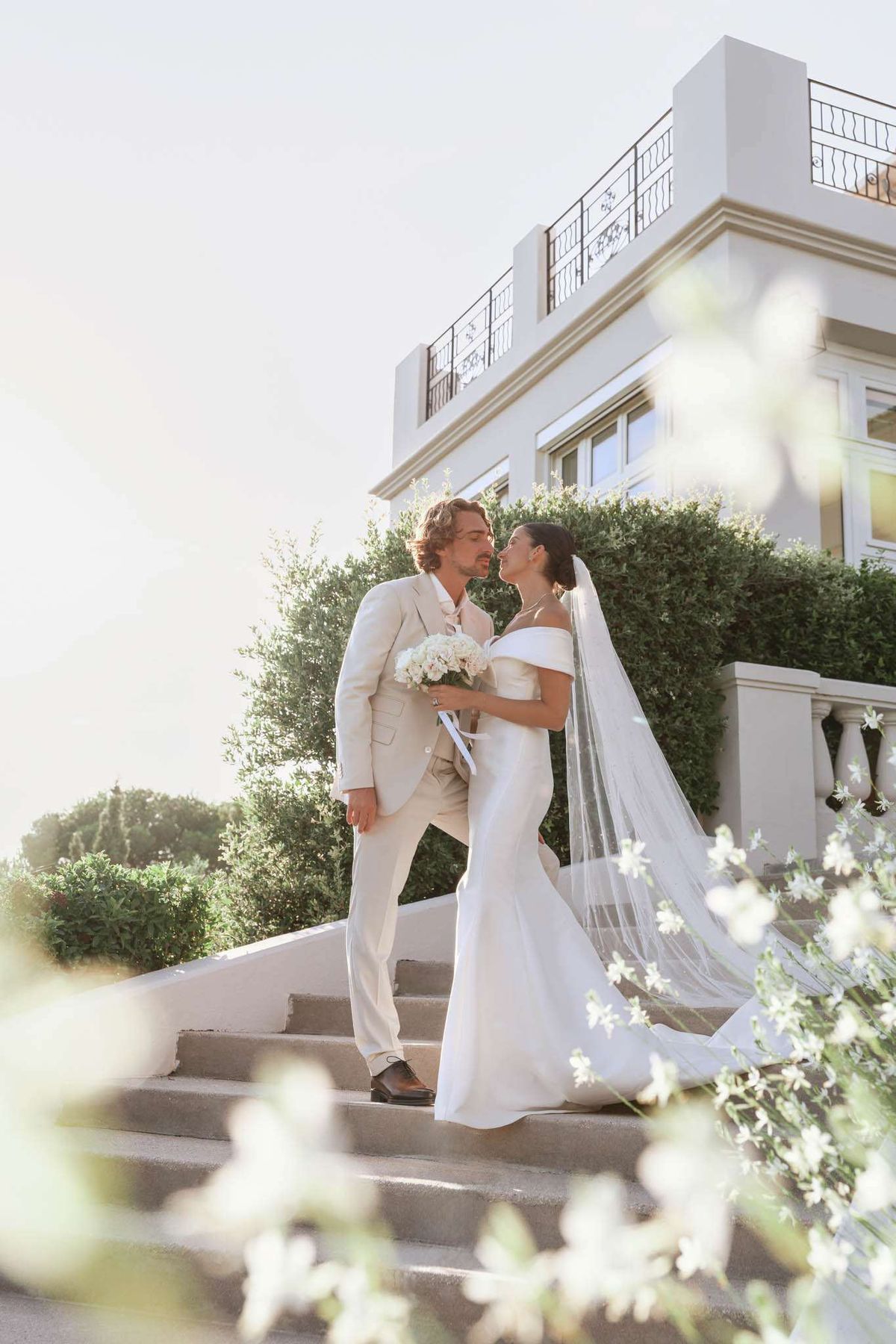 Couple about to kiss on villa staircase with bride holding white peony bouquet and cathedral veil