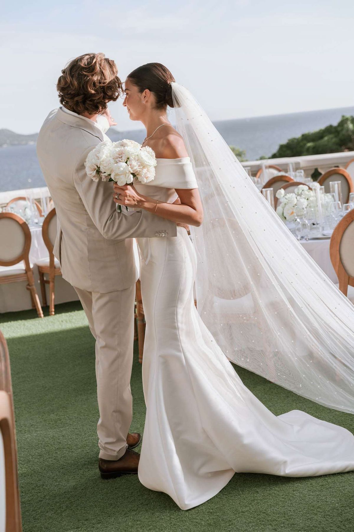 Couple embraces on waterfront terrace with pearl-embellished veil and white peony bouquet at reception setup